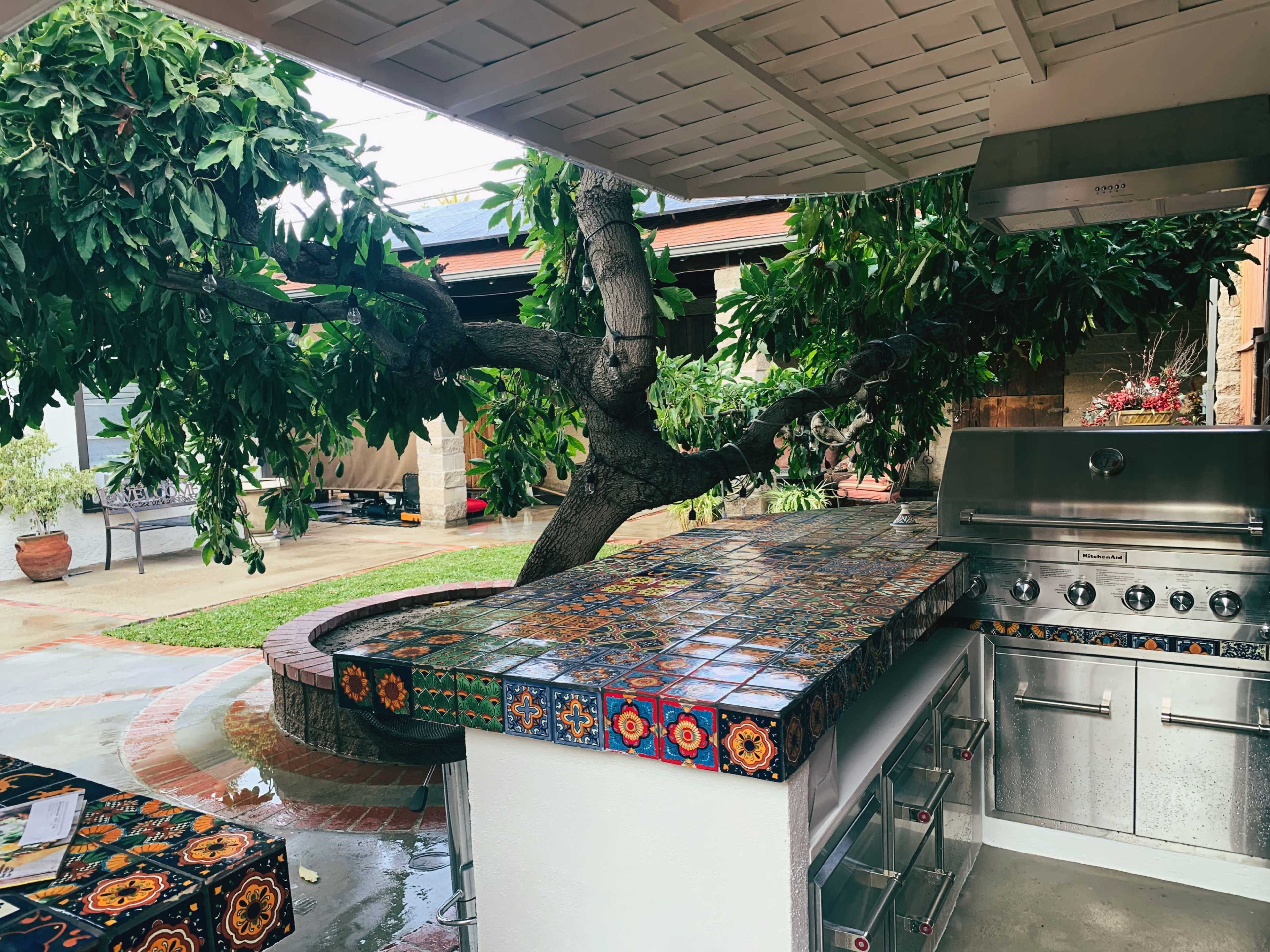 An outdoor kitchen features a grilling area beneath a large tree, with a colorful tiled countertop and metal storage drawers.
