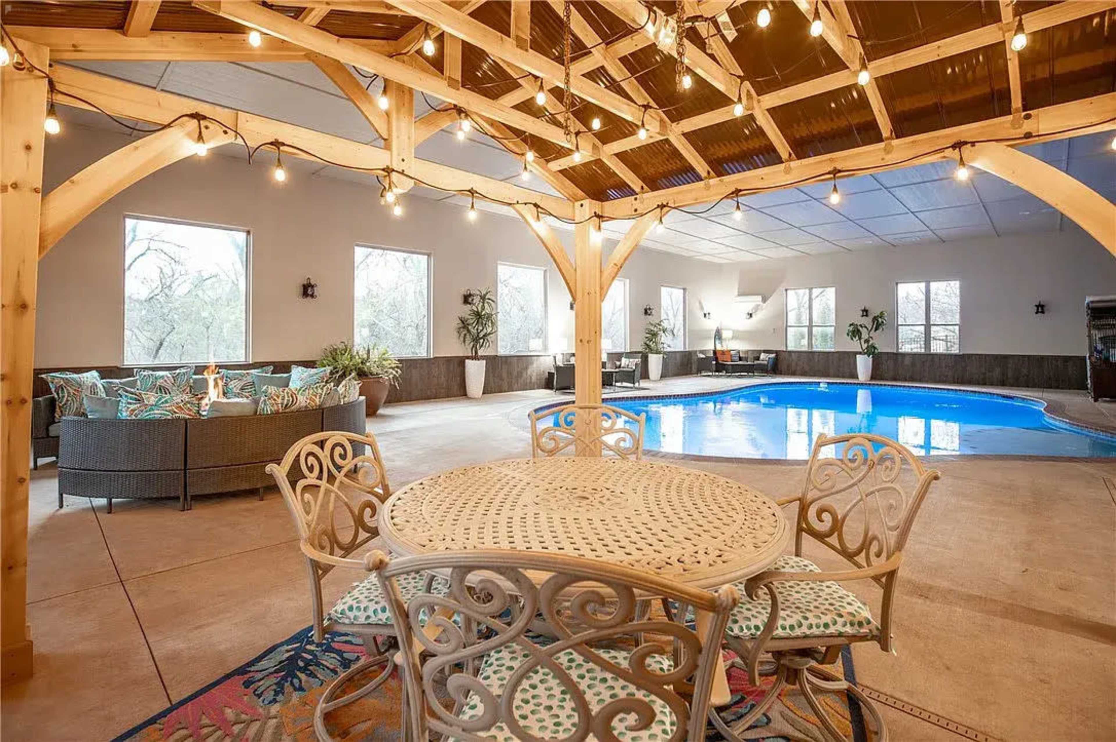 An indoor area with a wooden structure that includes a circular table surrounded by chairs, a cozy seating arrangement, and a swimming pool in the background.