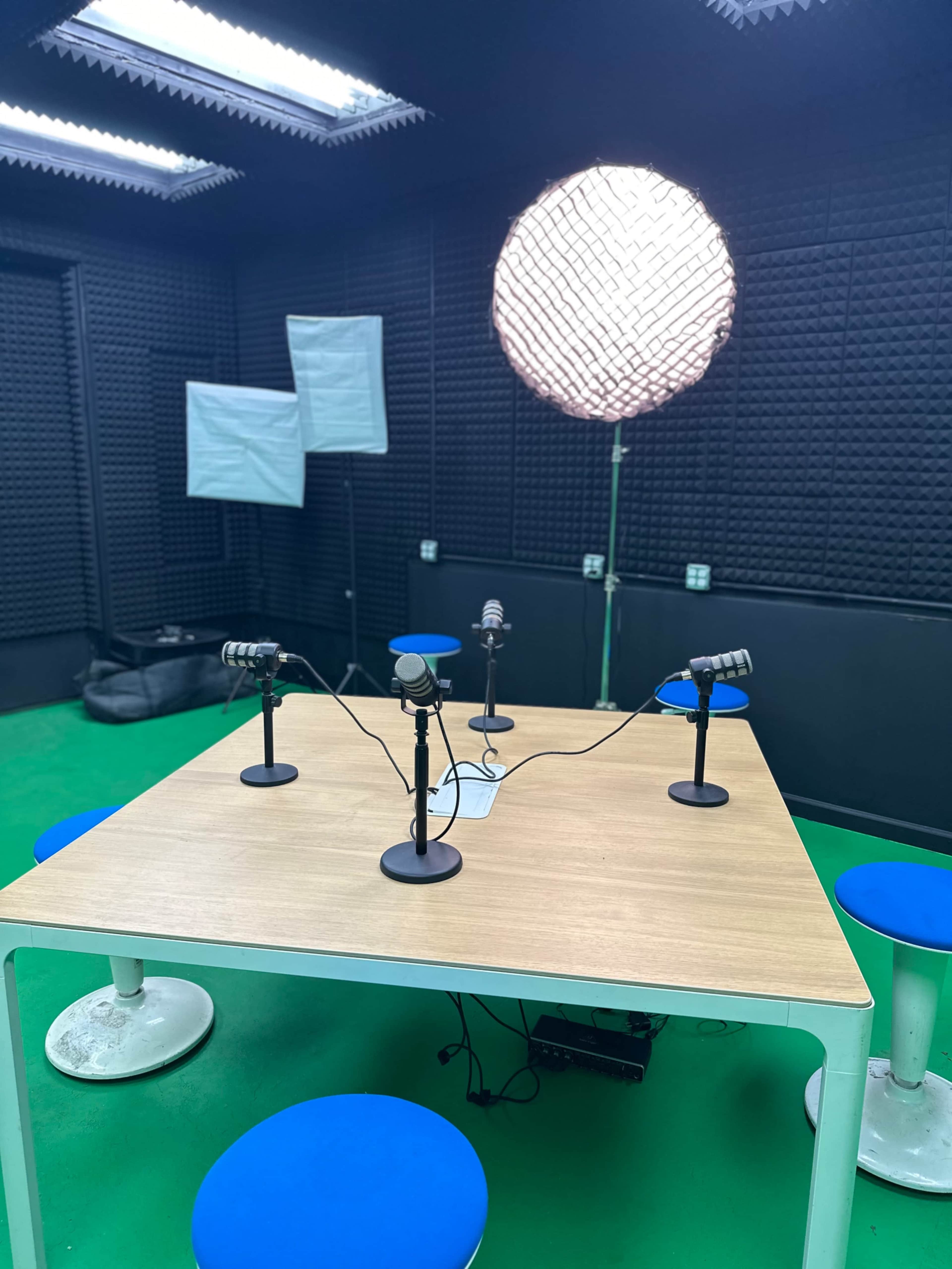 The image shows a podcast studio featuring a wooden table surrounded by blue stools, with several microphones positioned on the table and a large light source overhead.