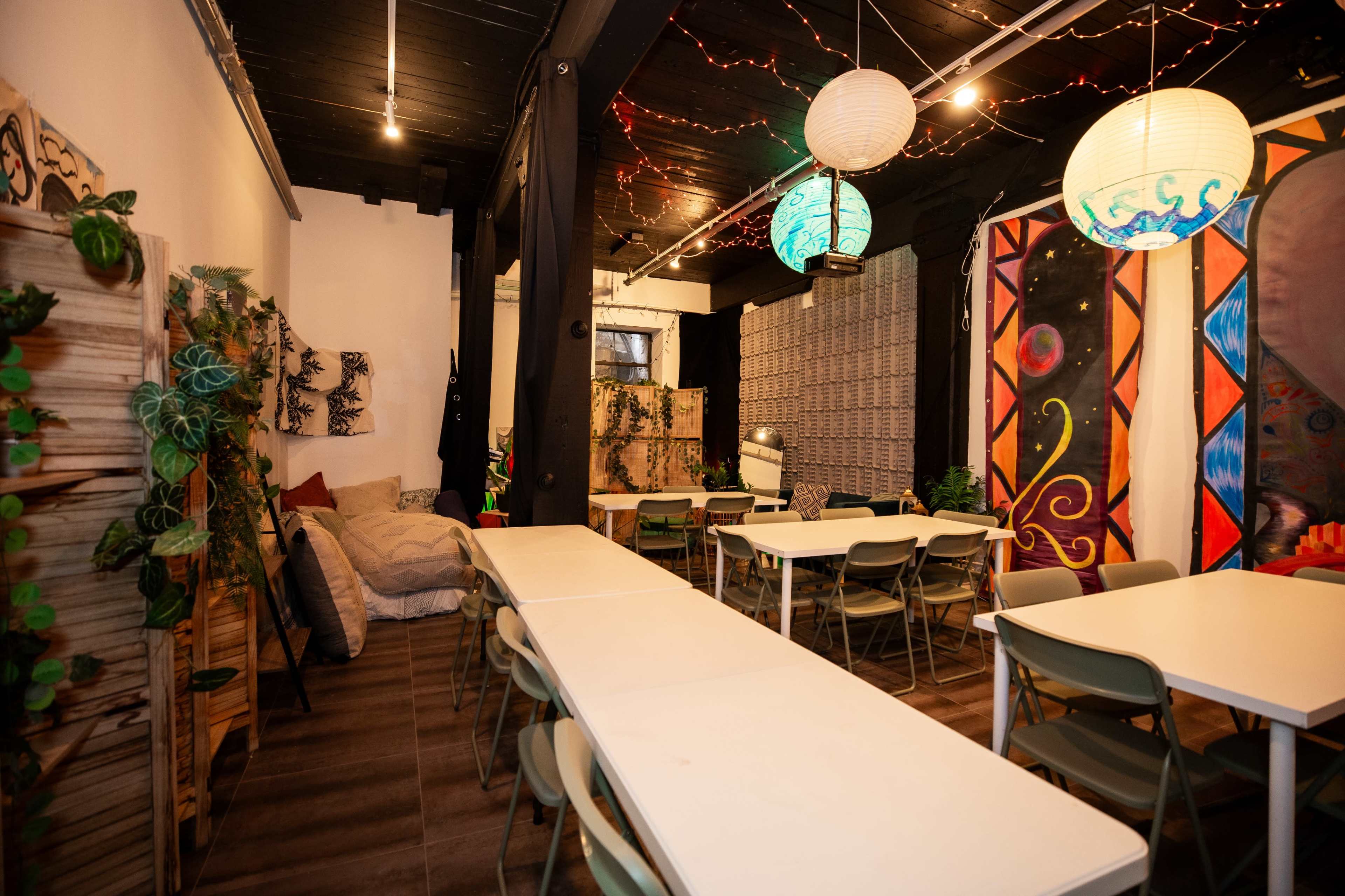 The image shows a brightly lit communal space featuring several long tables, chairs, and decorative wall art, with plants scattered throughout the room.