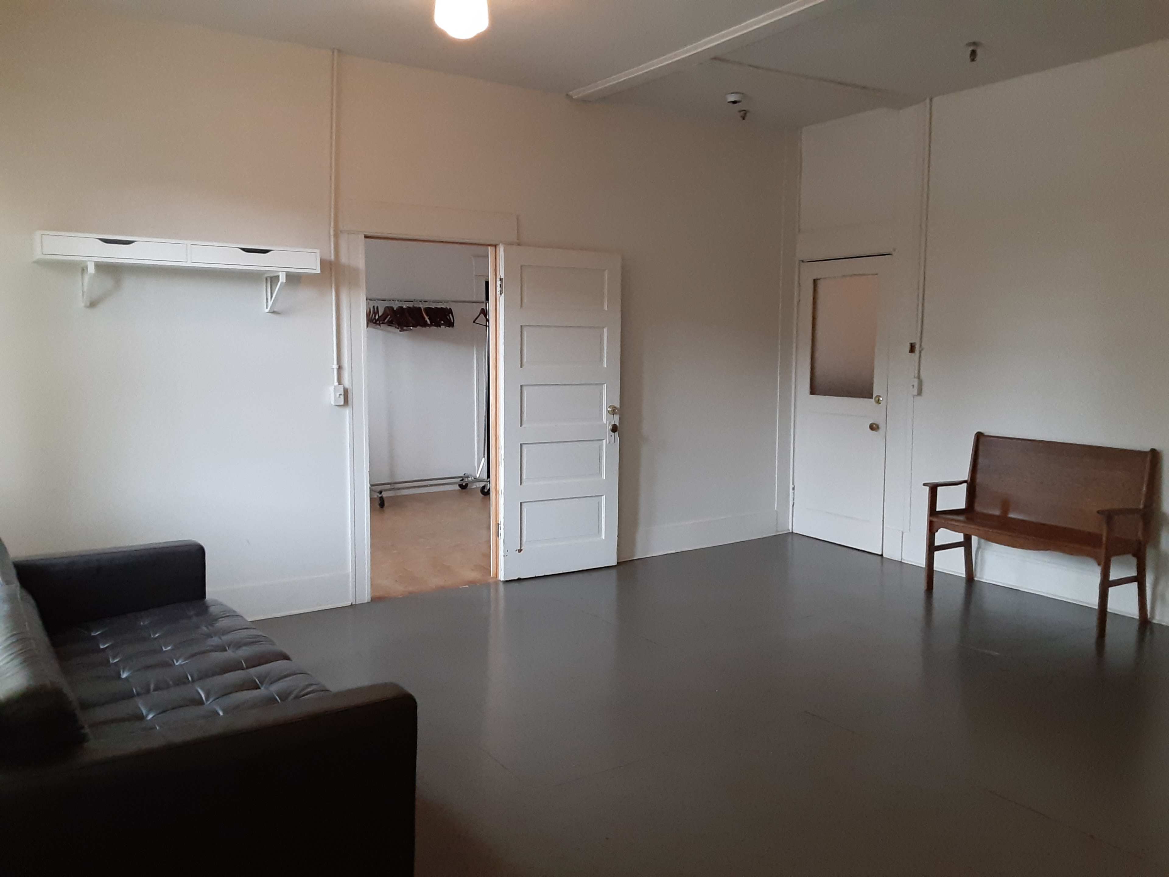 A sparsely furnished room with a black sofa on the left, a wooden bench on the right, and empty walls.