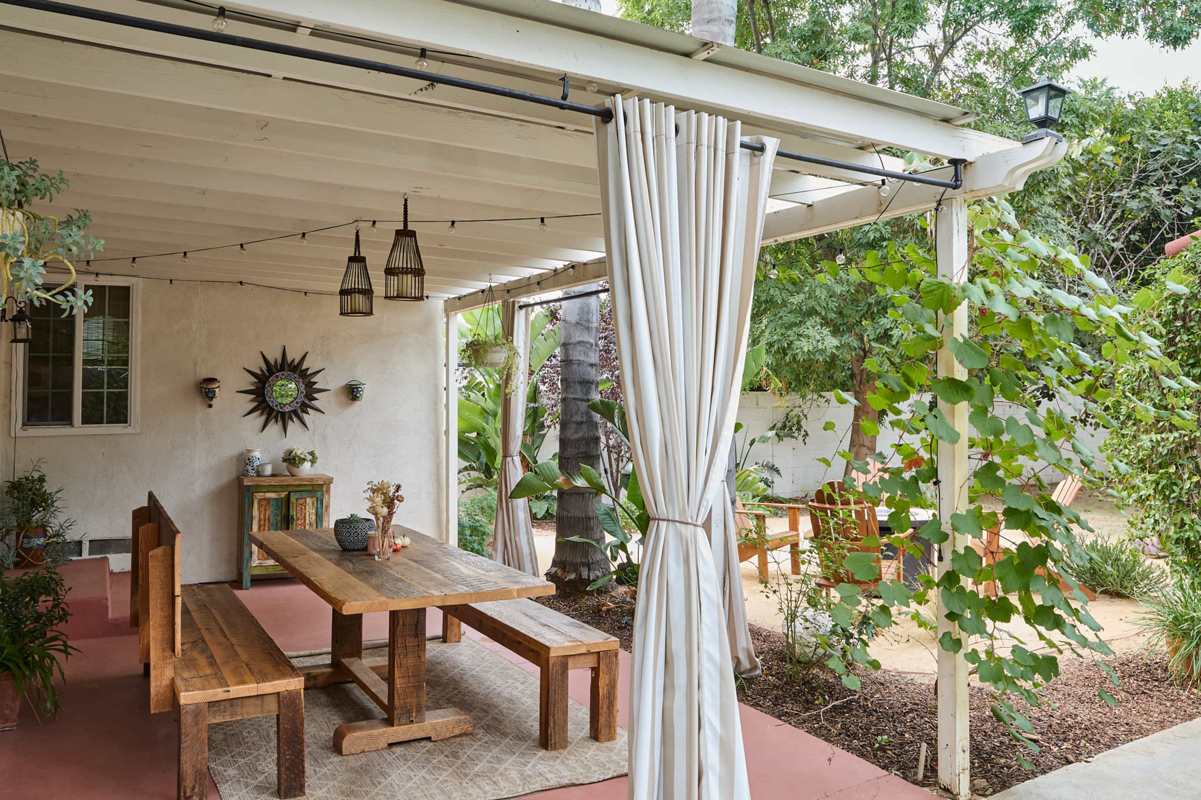 The image shows a covered outdoor seating area with a wooden table and benches, surrounded by greenery and pendant lights.