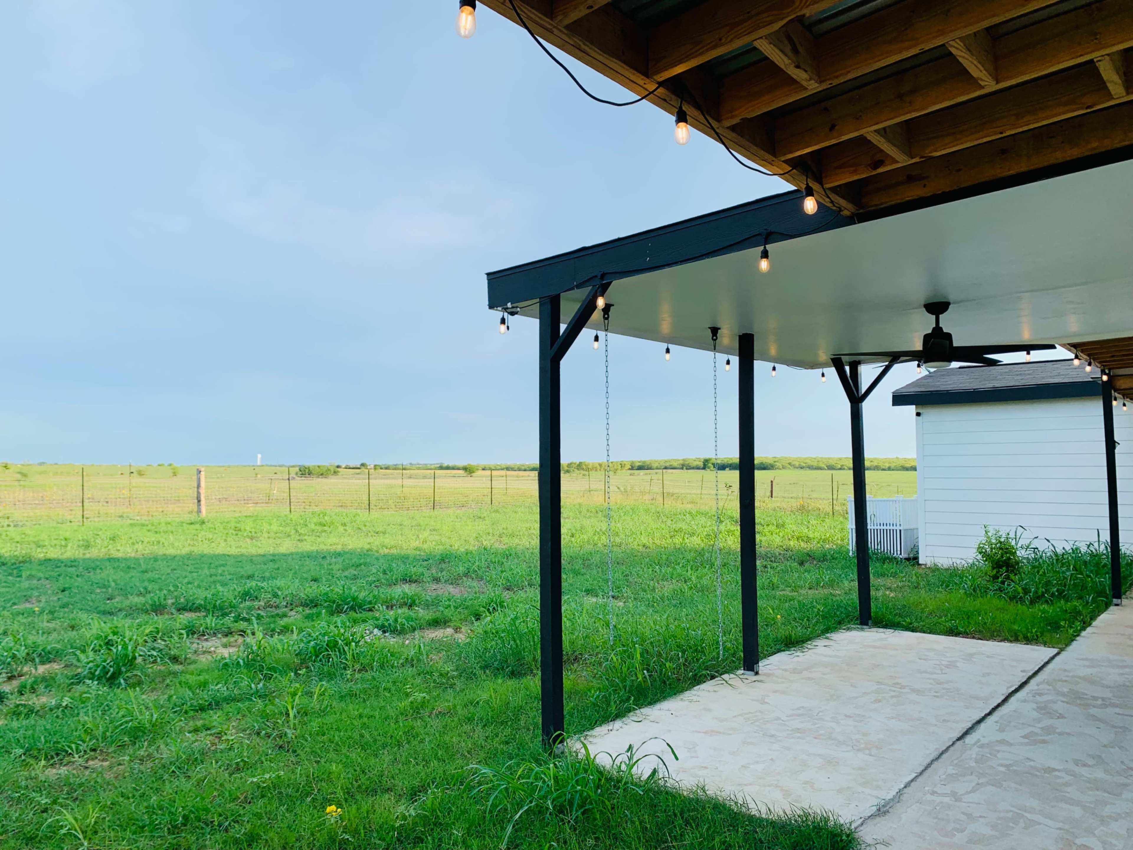 The image shows a covered patio with string lights overlooking a grassy field and a clear sky.