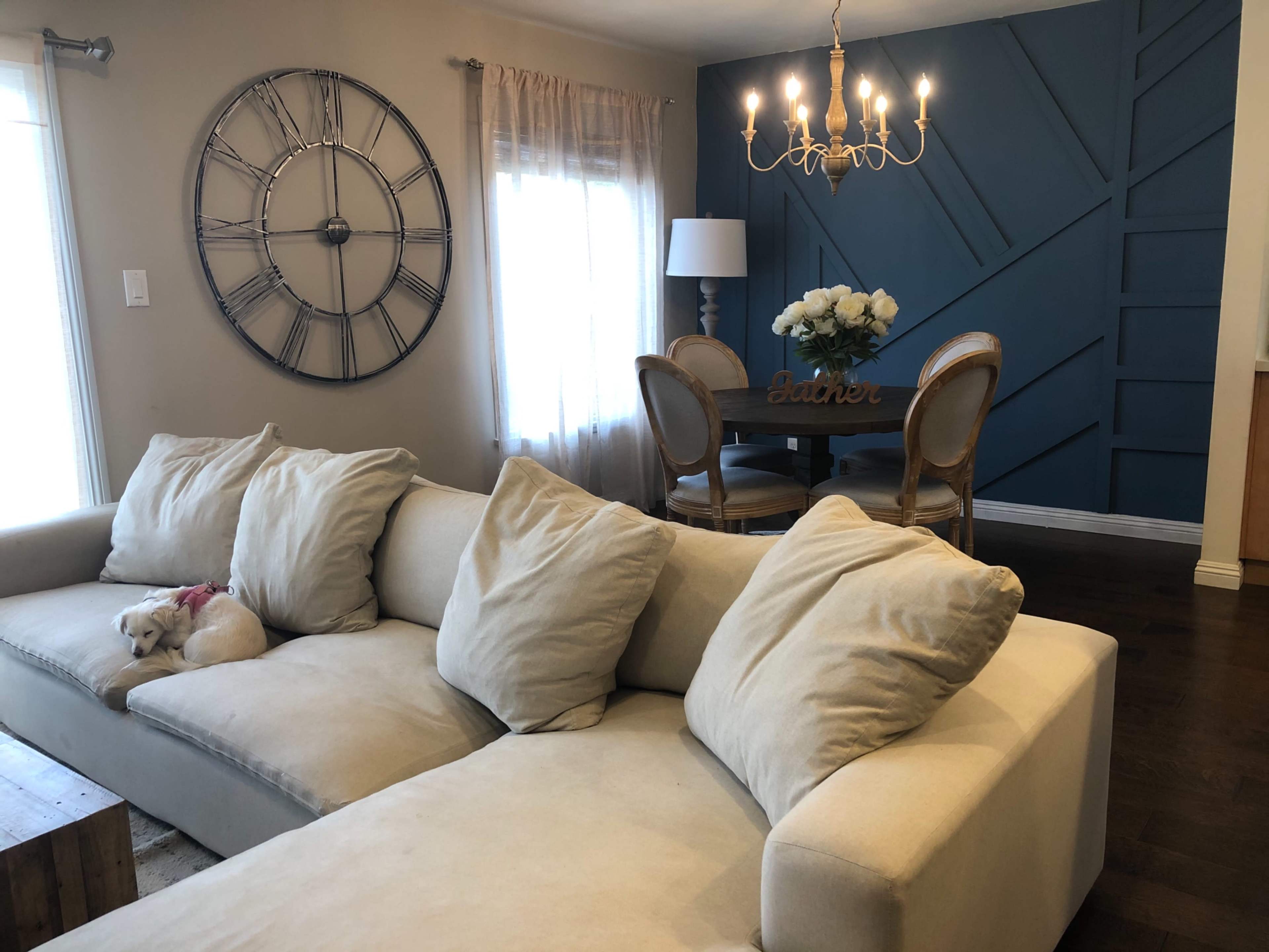 A light-colored sectional sofa is positioned in a cozy living room featuring a large wall clock, a dining table with floral decor, and a chandelier.
