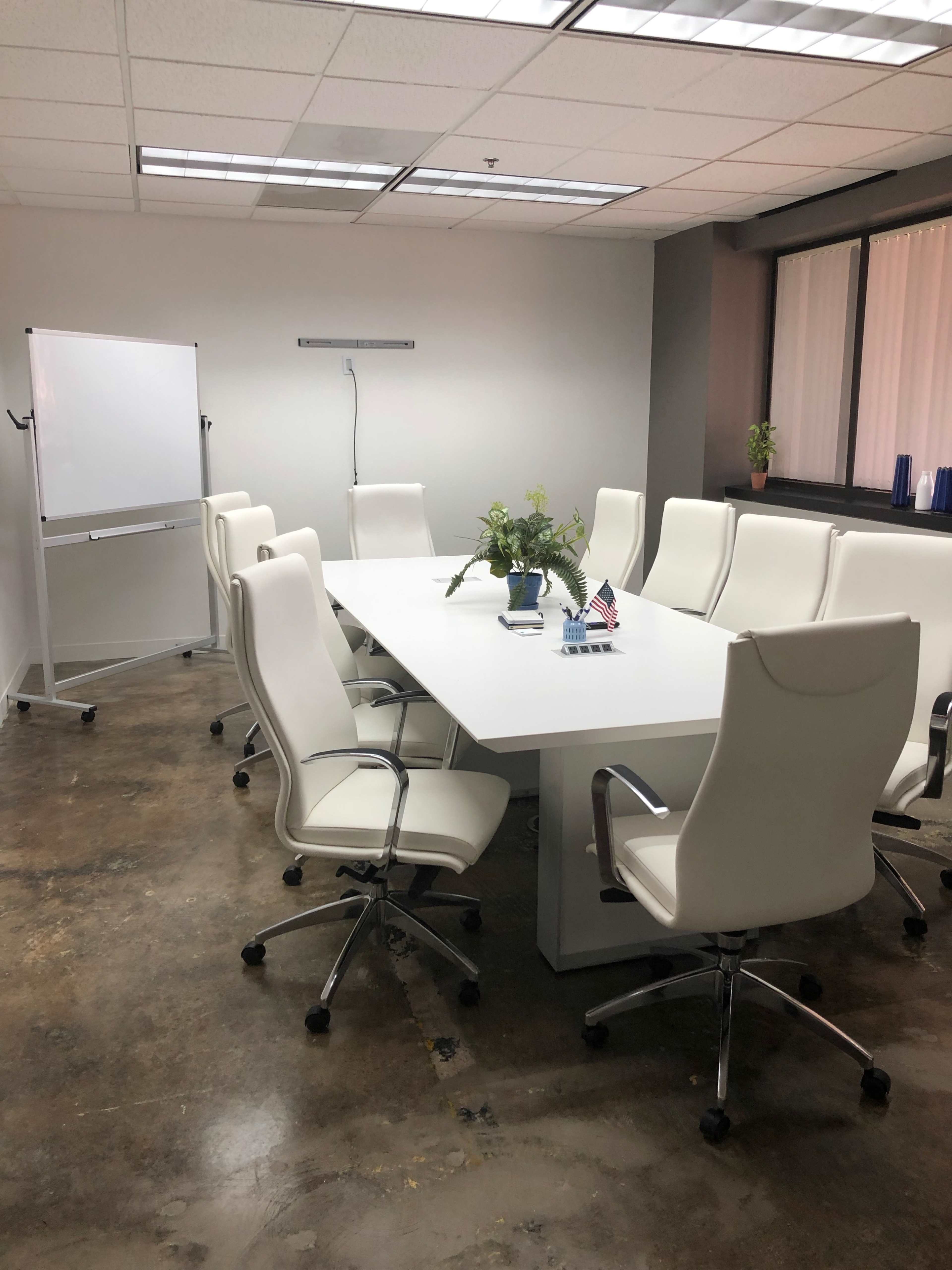 A conference room features a long white table surrounded by eight white chairs, with a flip chart and a small potted plant on the table.