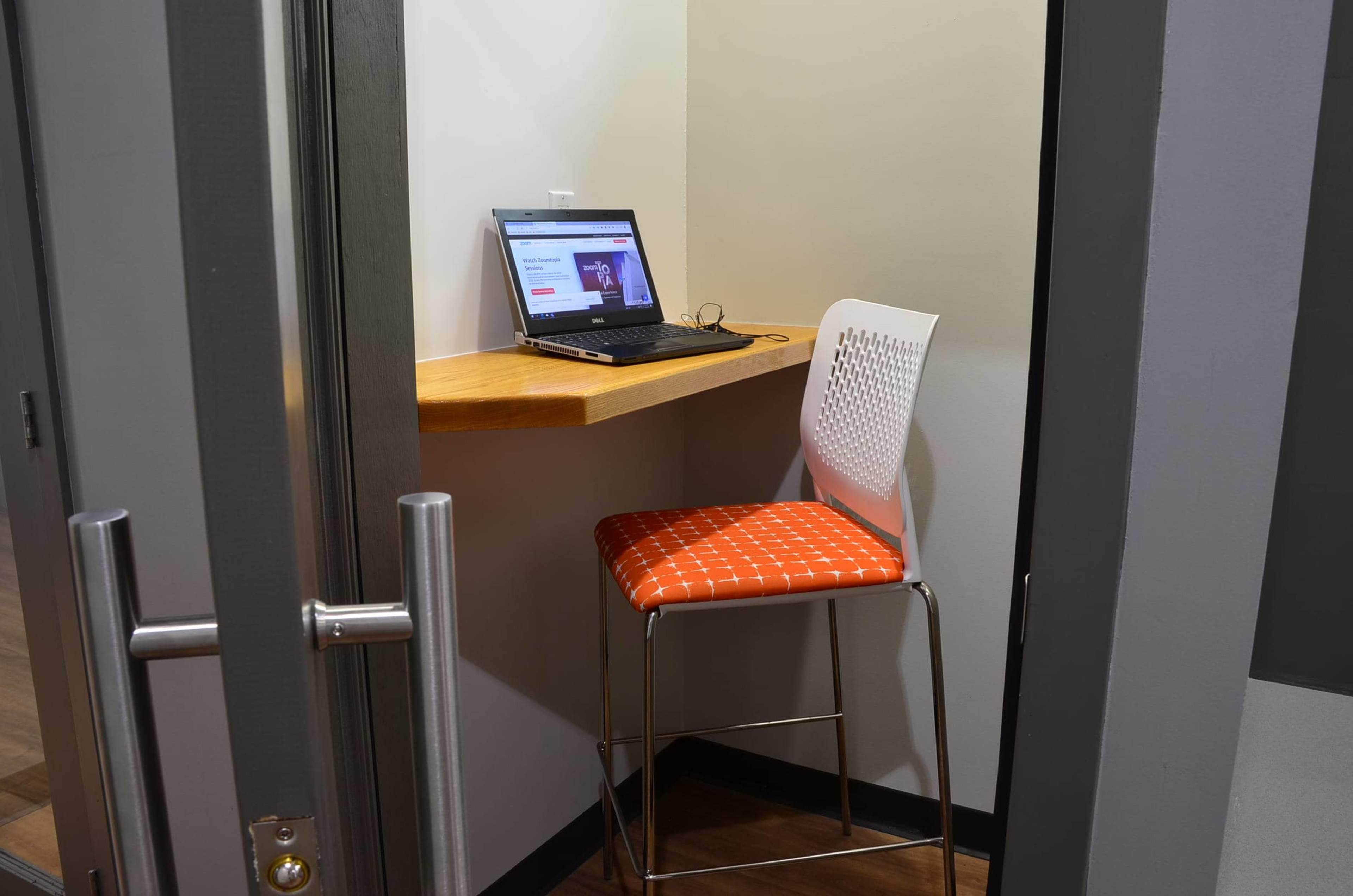 The image shows a small workspace featuring a laptop on a wooden ledge next to a high stool with an orange patterned cushion, located in a corner of a room.