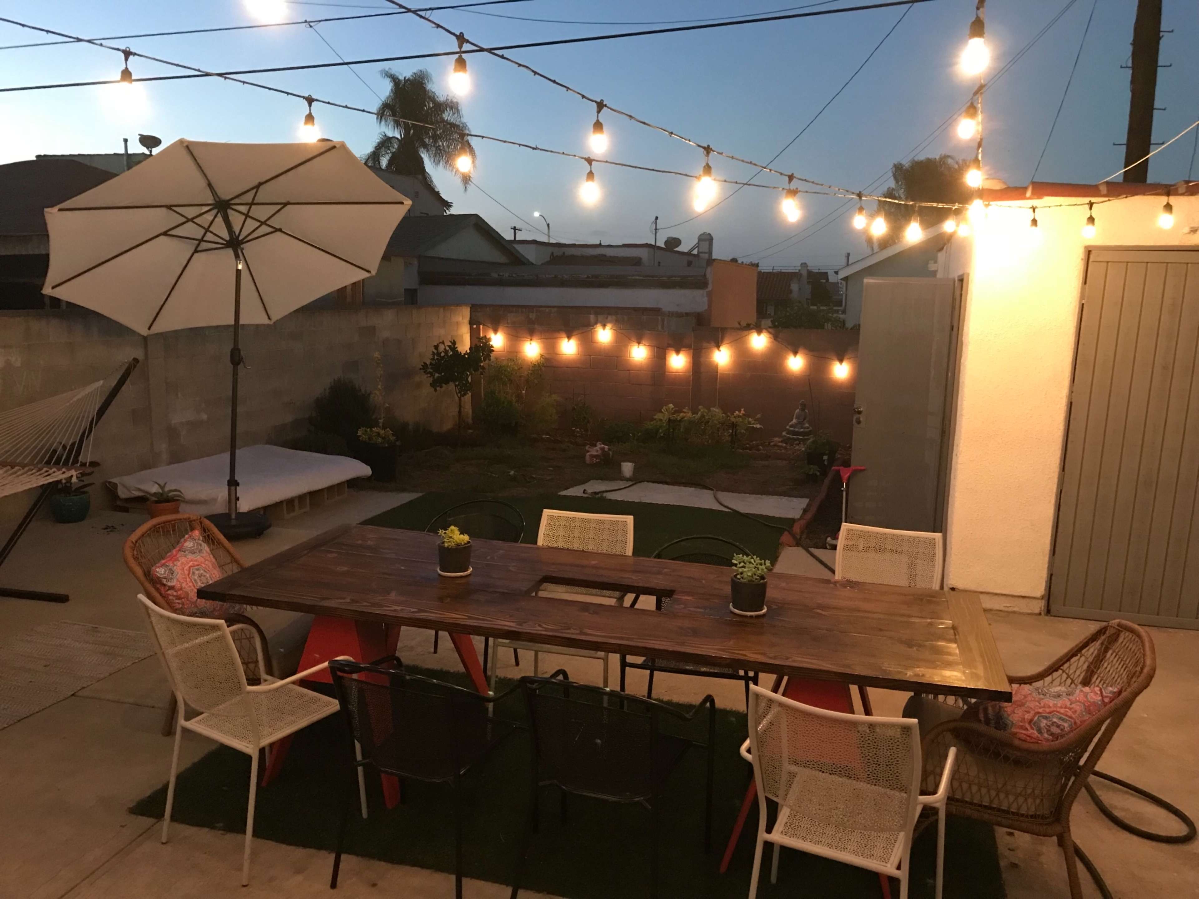 A wooden dining table surrounded by six chairs is illuminated by string lights in a backyard setting with a large umbrella and a hammock in the background.