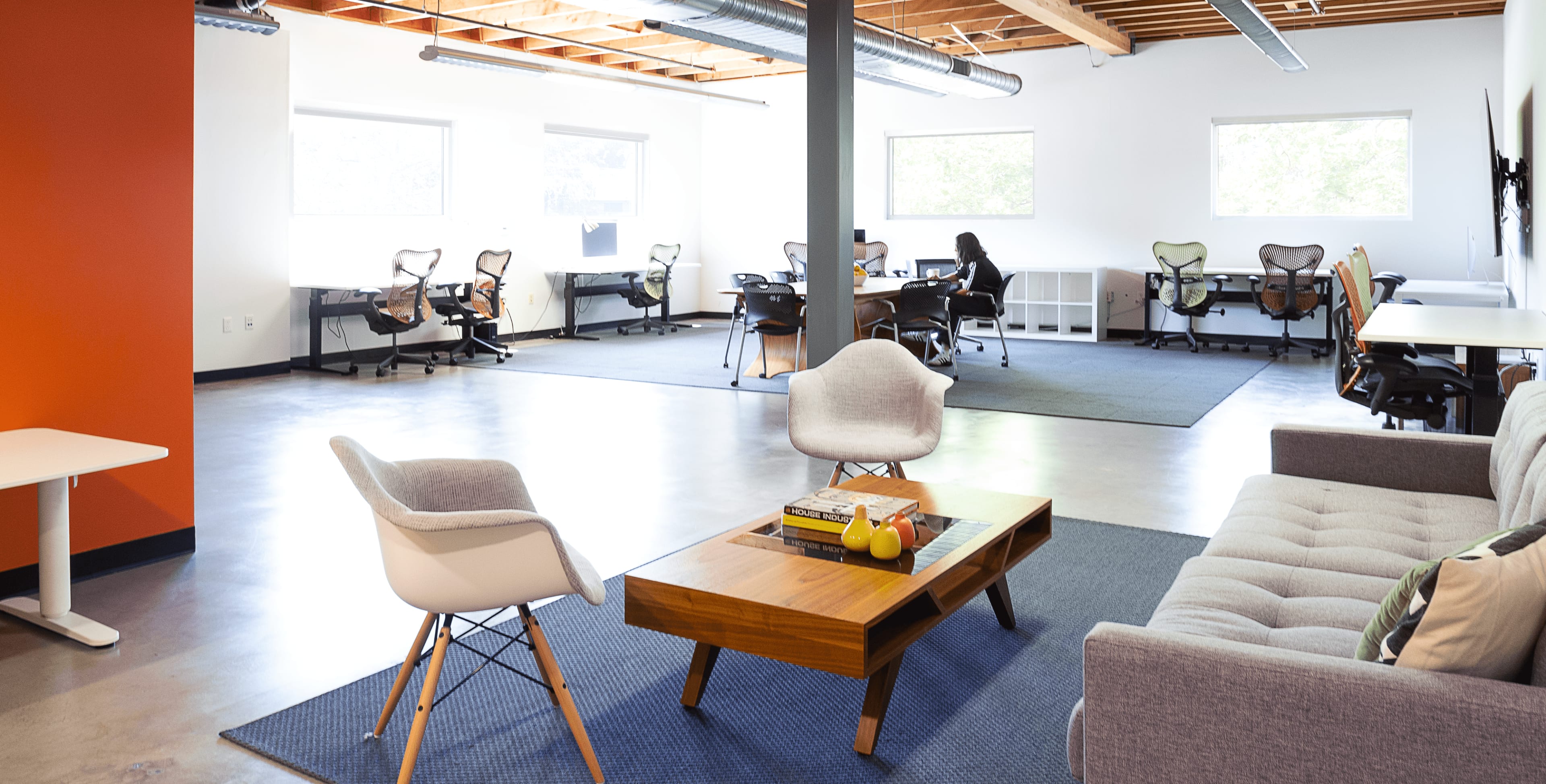 A modern office space with several workstations, a seating area with a coffee table, and large windows providing natural light.