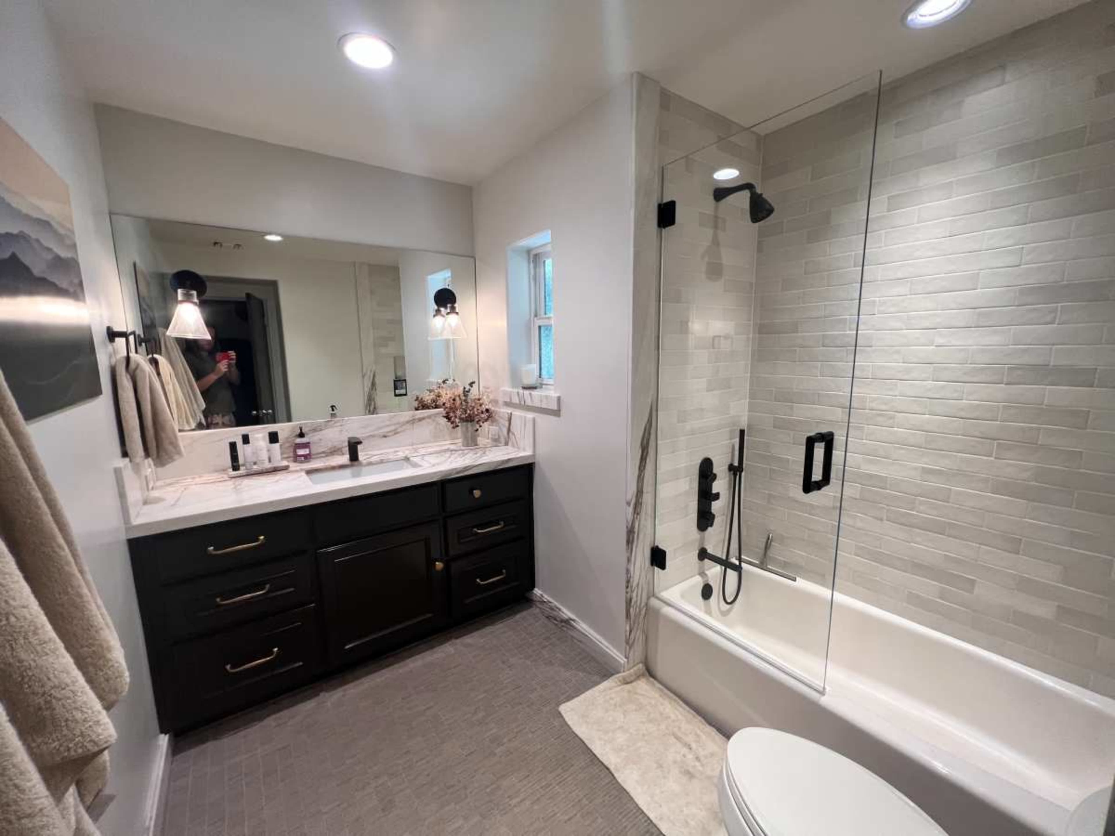 The image shows a modern bathroom featuring a glass shower, a bathtub, a double vanity with dark cabinetry, and neutral tile flooring.