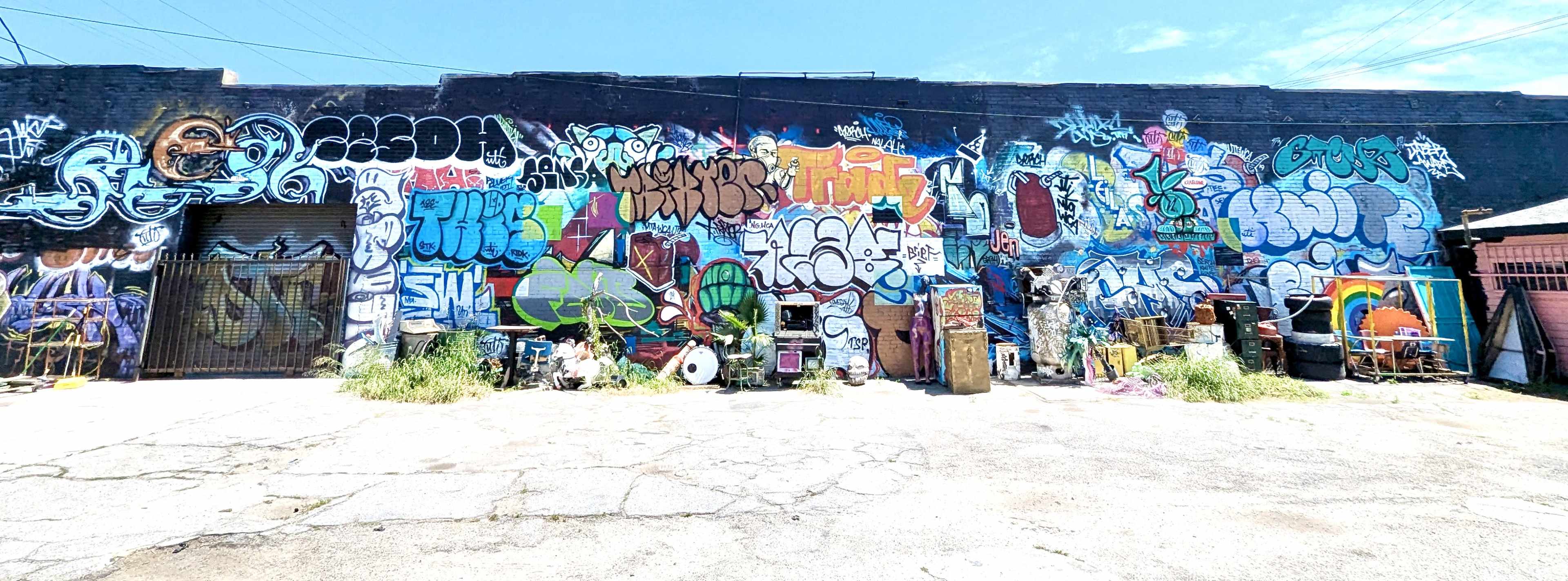 The image shows a large brick wall covered in vibrant graffiti art, with an assortment of discarded furniture and items in the foreground.