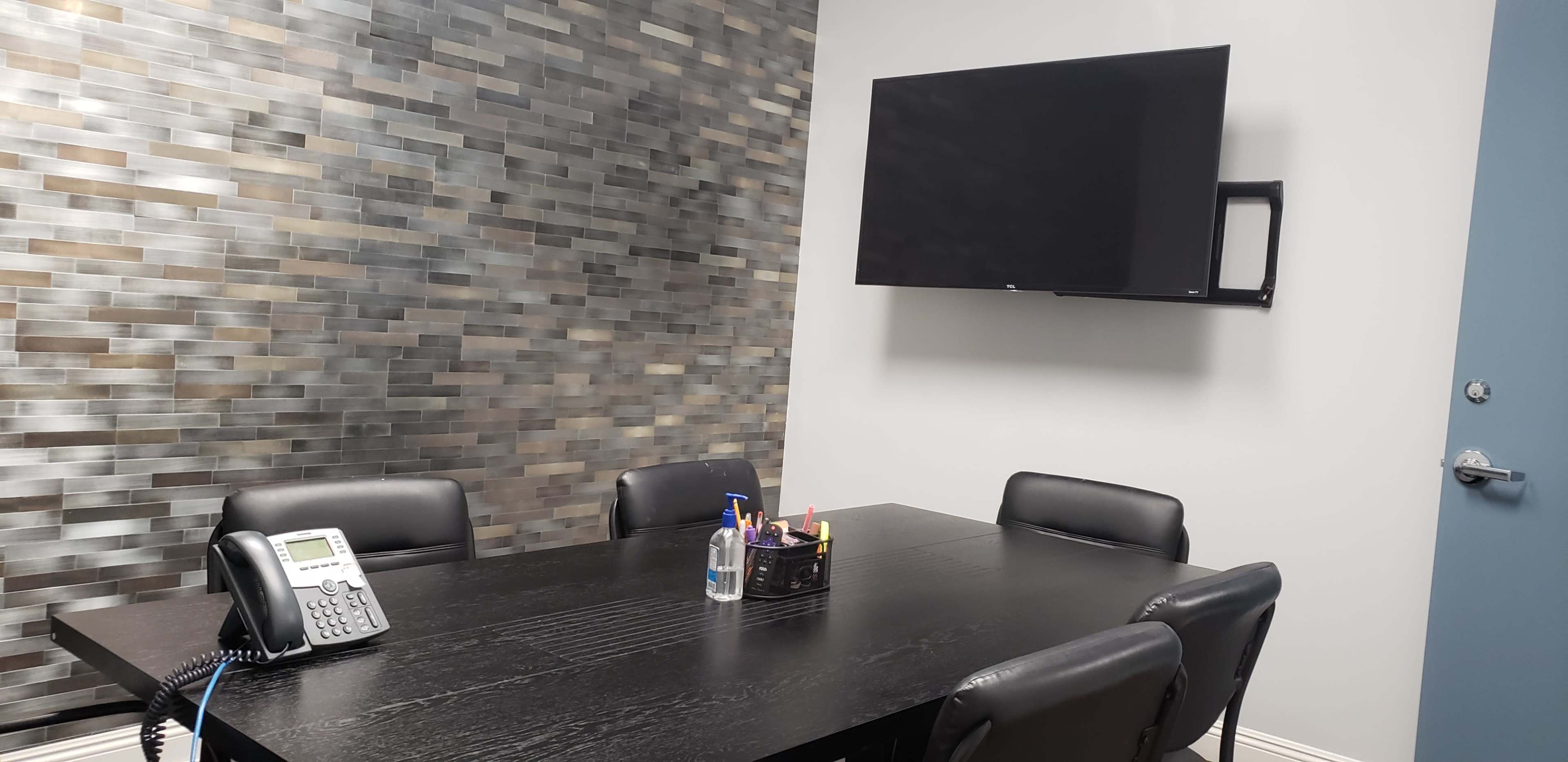 A conference room features a black table surrounded by chairs, a wall-mounted television, and a stone-tiled accent wall.