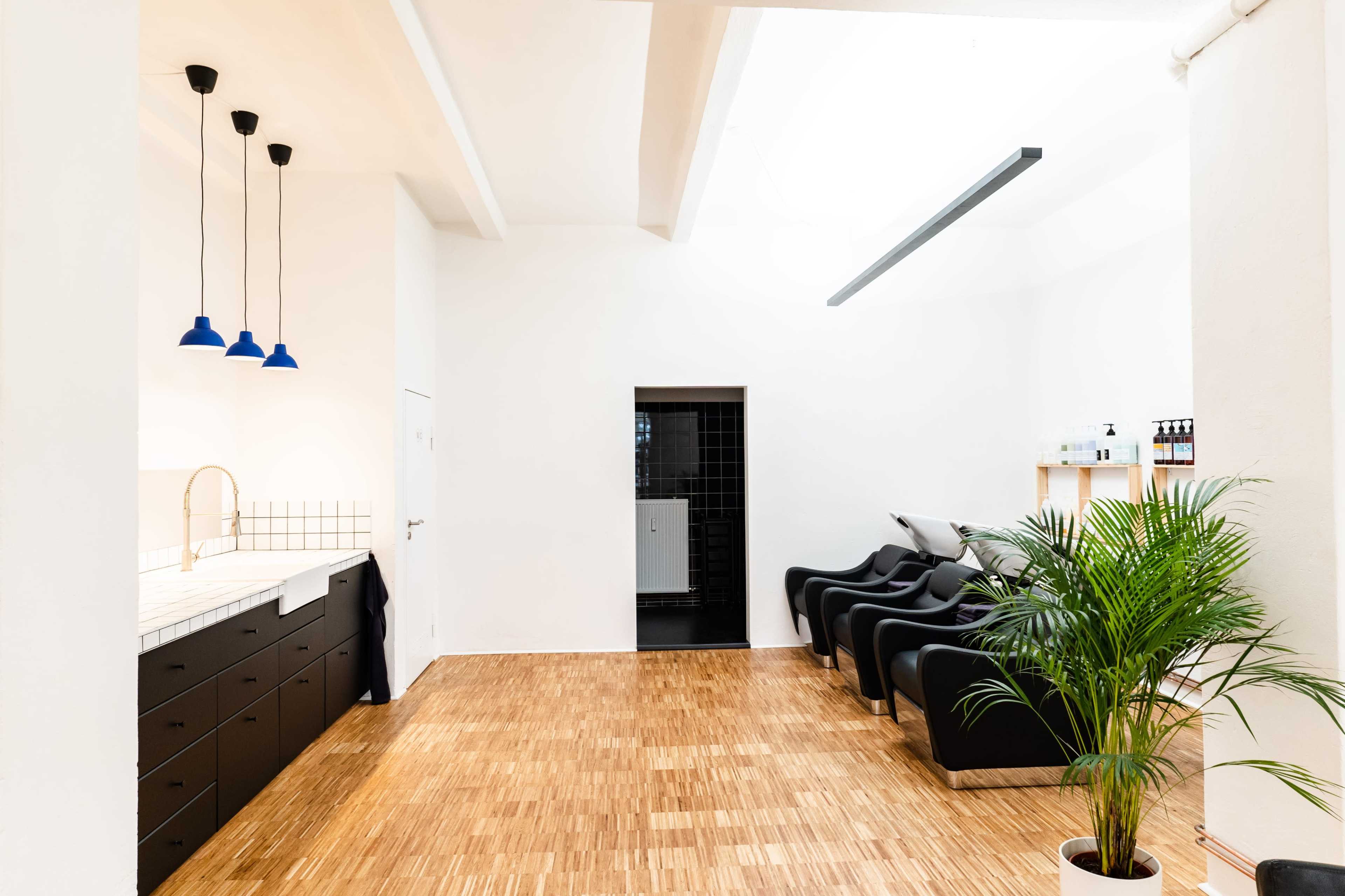 A spacious room with a wooden floor, featuring a counter with black drawers, three blue pendant lights, black chairs, and a potted plant in one corner.