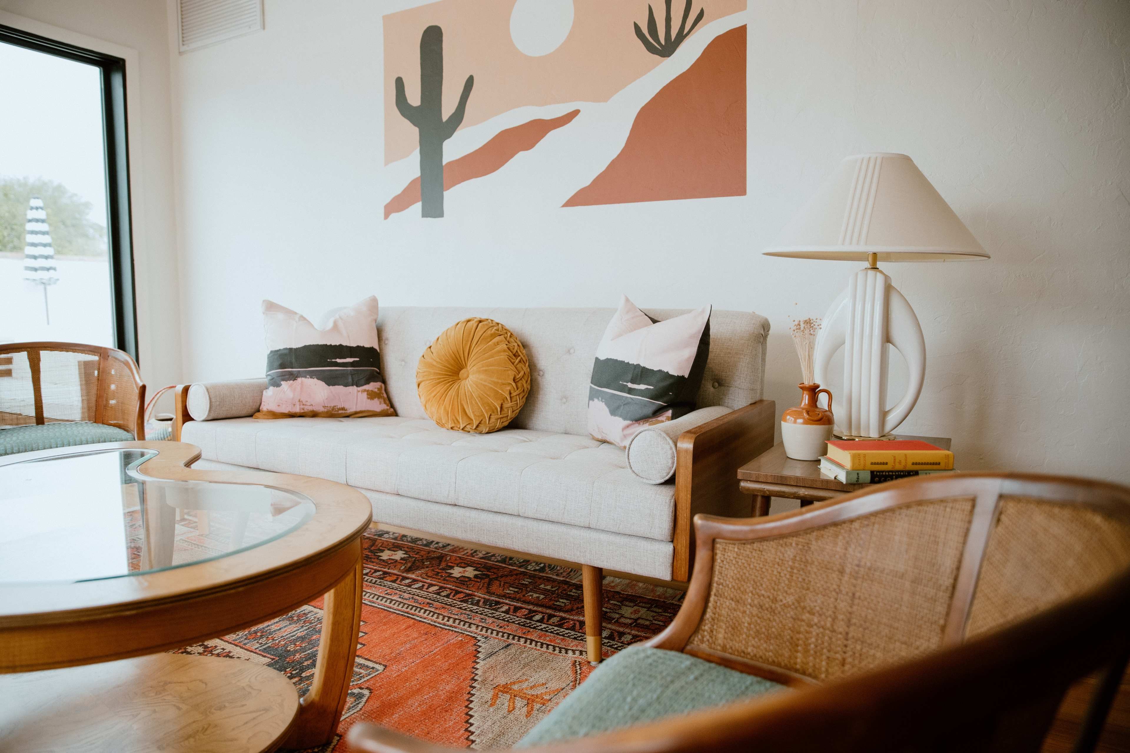 A modern living room features a light-colored sofa with decorative pillows, a round orange cushion, a mid-century lamp, and a patterned rug.