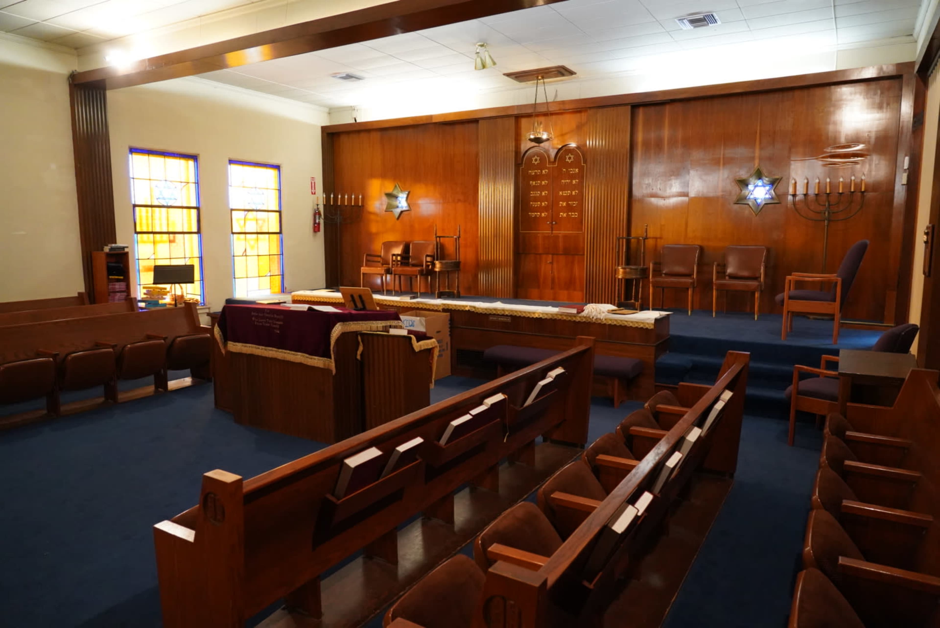 The interior of a synagogue features wooden benches, a central raised platform with a Torah ark, and colorful stained-glass windows.