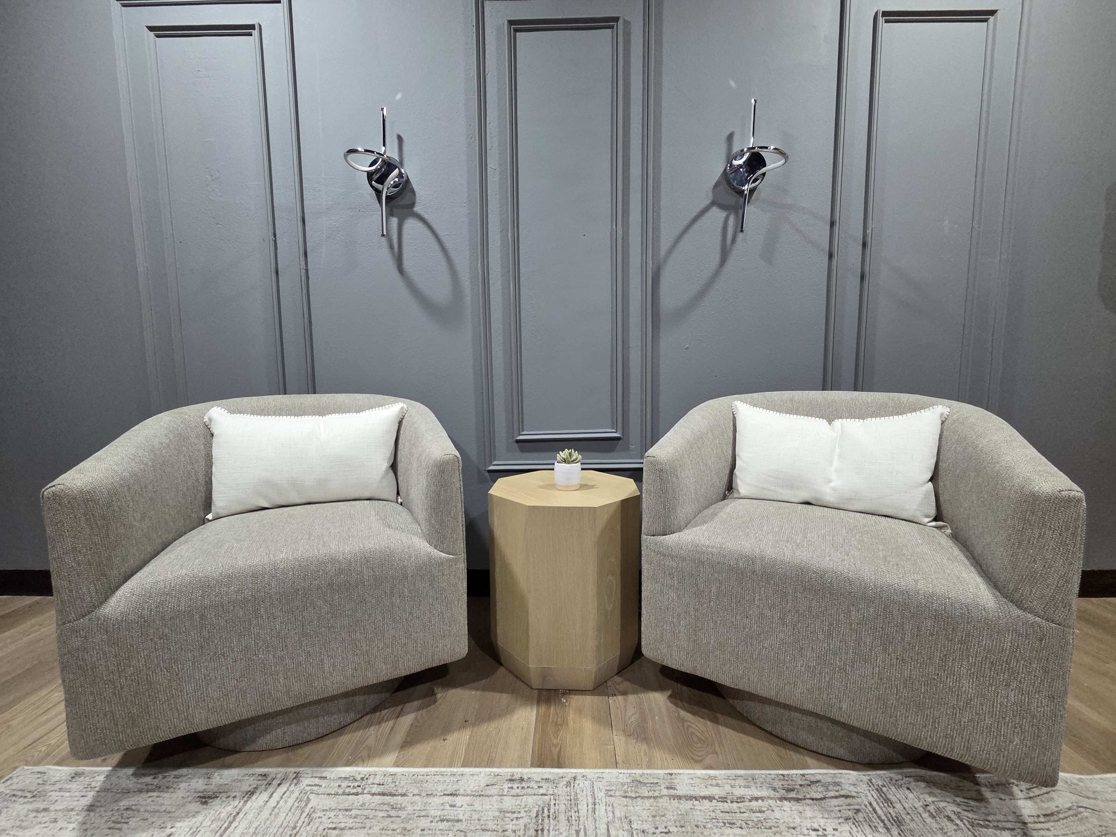 The image features two modern, gray upholstered chairs with white throw pillows positioned opposite each other, accompanied by a small wooden side table in a minimalist interior setting.