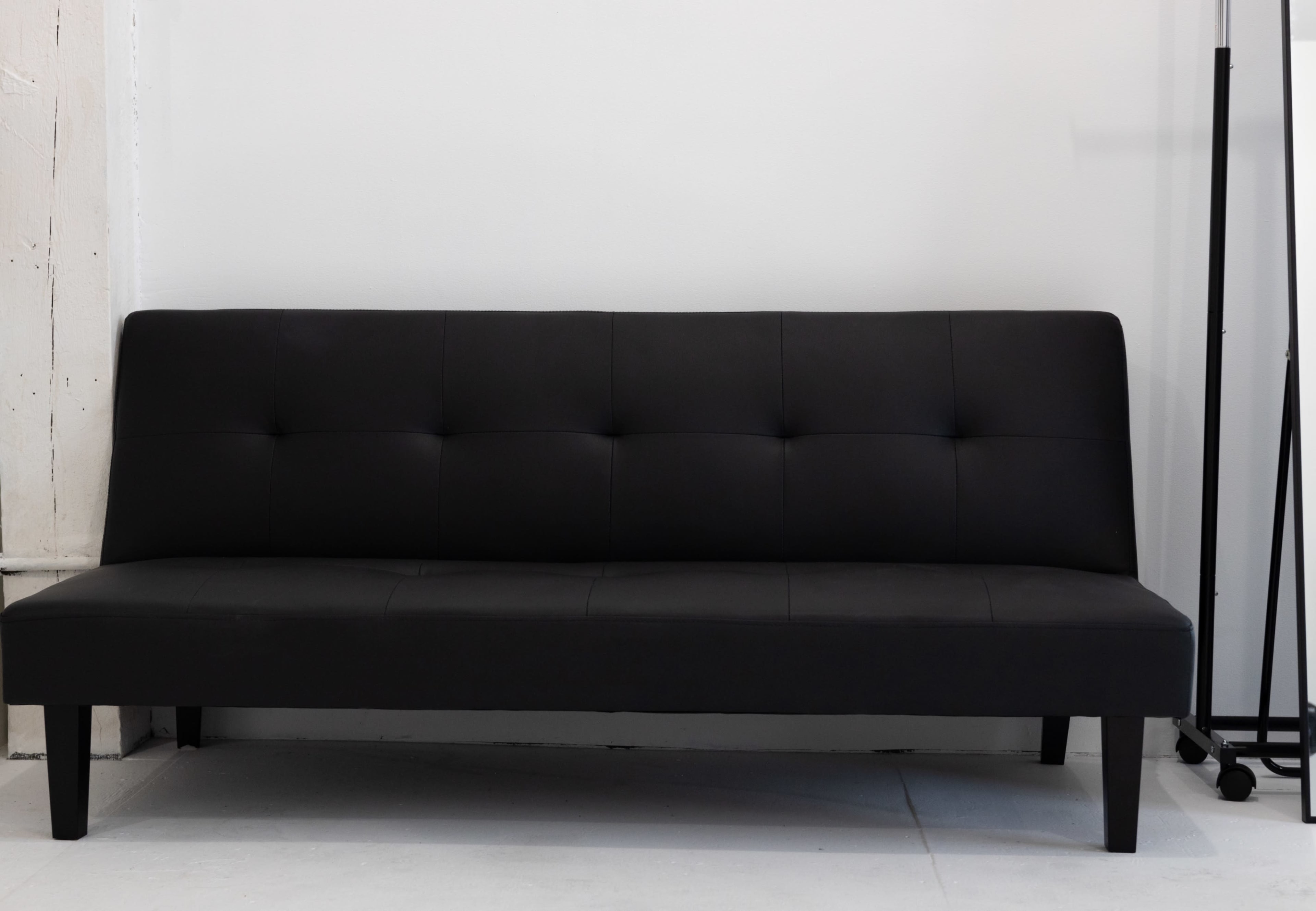 The image shows a black, upholstered sofa with a minimalist design, positioned against a plain white wall.