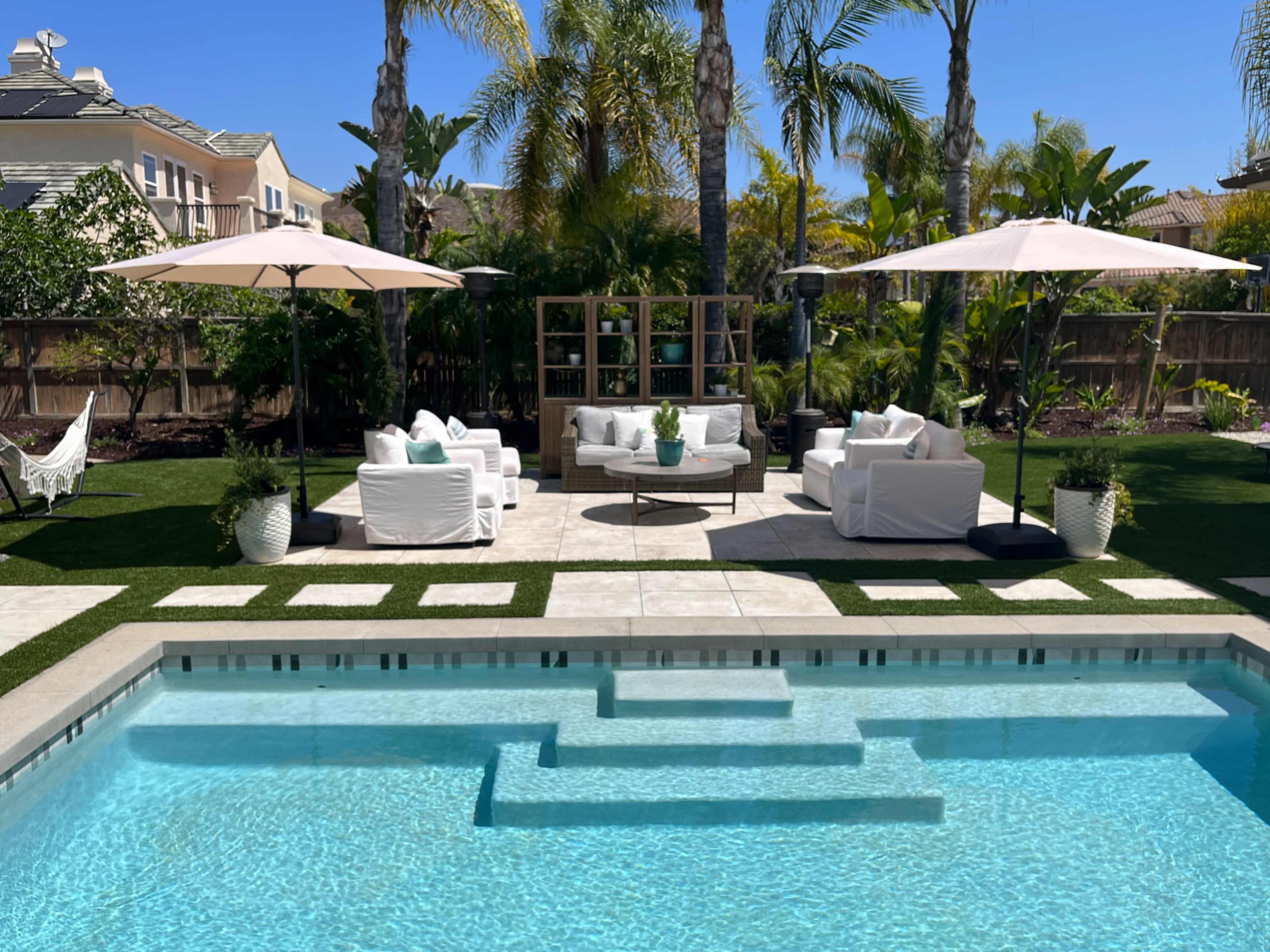 A landscaped backyard features a swimming pool with stepped entry, white outdoor furniture under umbrellas, and palm trees surrounding a seating area.