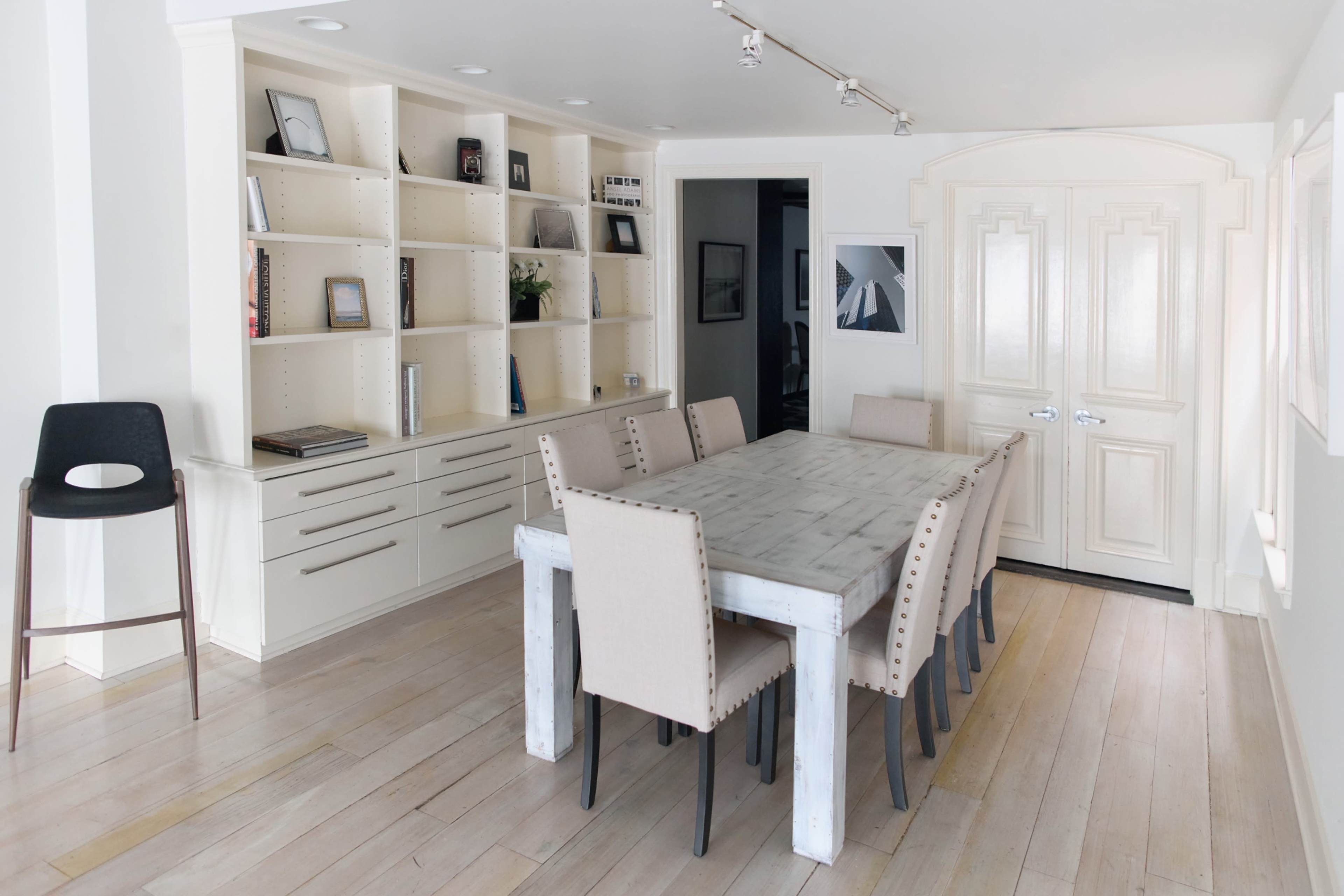 The image shows a modern dining area with a long wooden table and upholstered chairs, adjacent to a white bookshelf and an entry door.