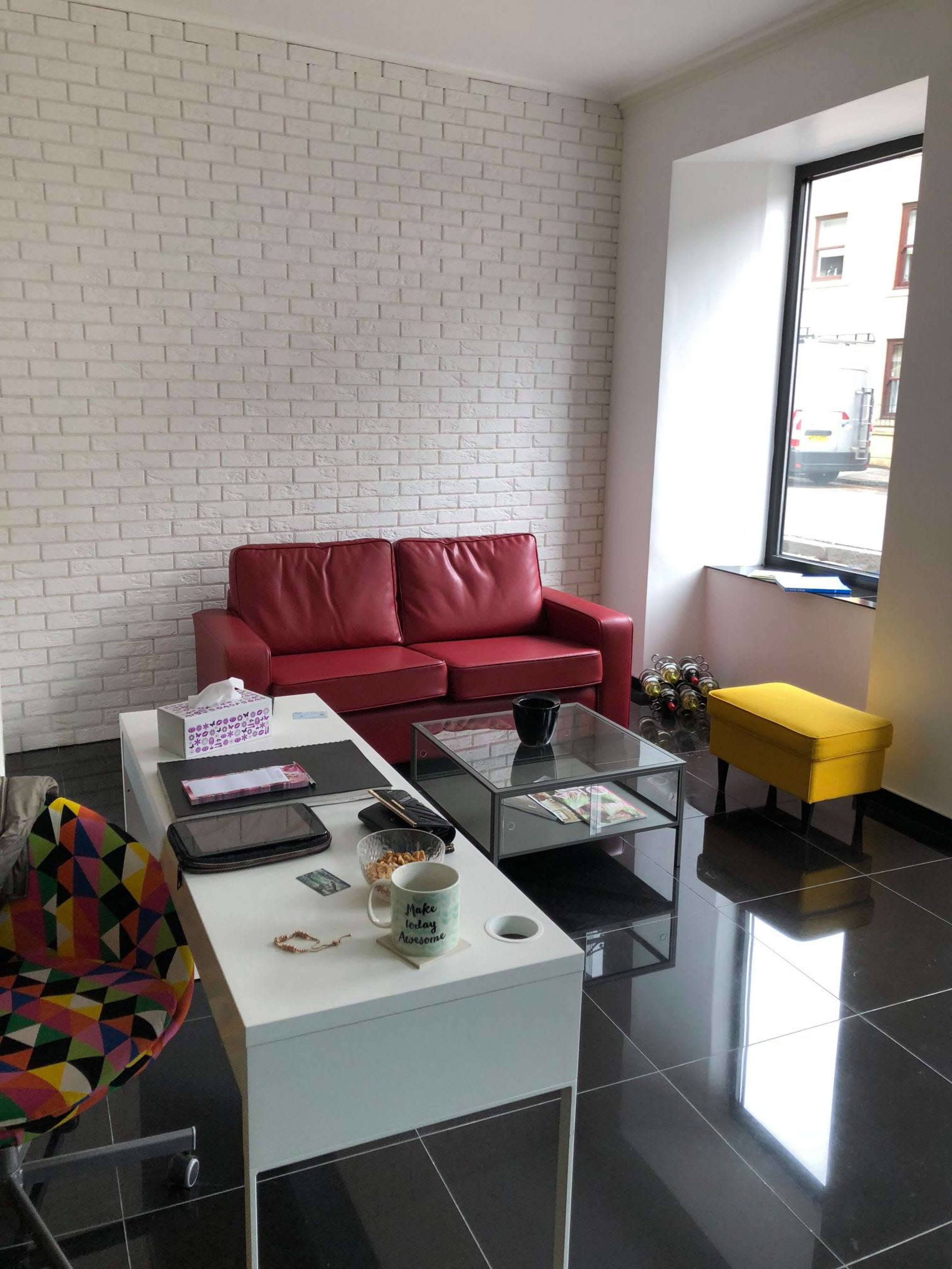 A modern living space with a red sofa, a yellow accent chair, and a glass coffee table, set against a white brick wall and black tiled floor.