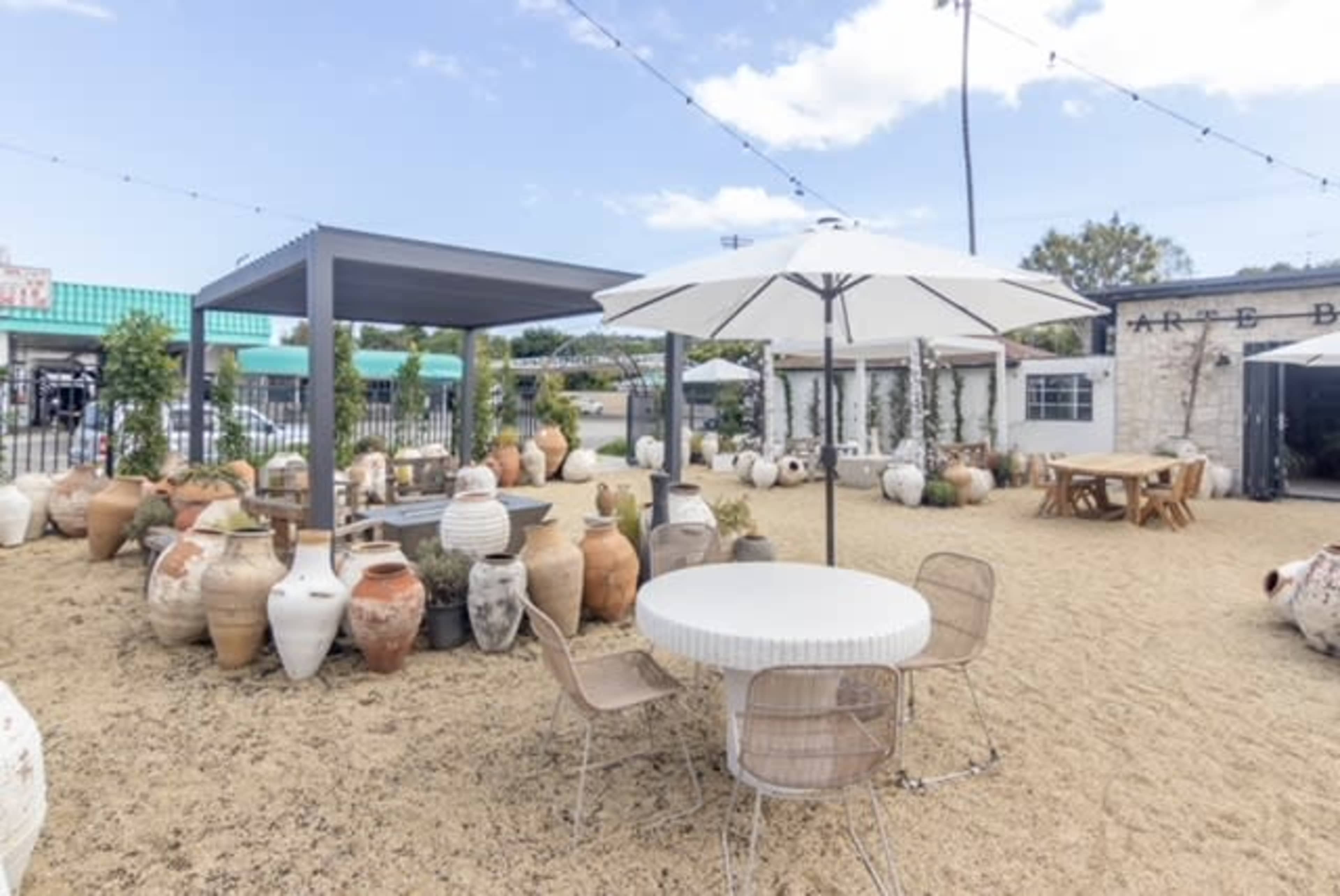 A courtyard features a circular white table surrounded by metal chairs, with numerous large pottery jars scattered on the sandy ground and string lights overhead.