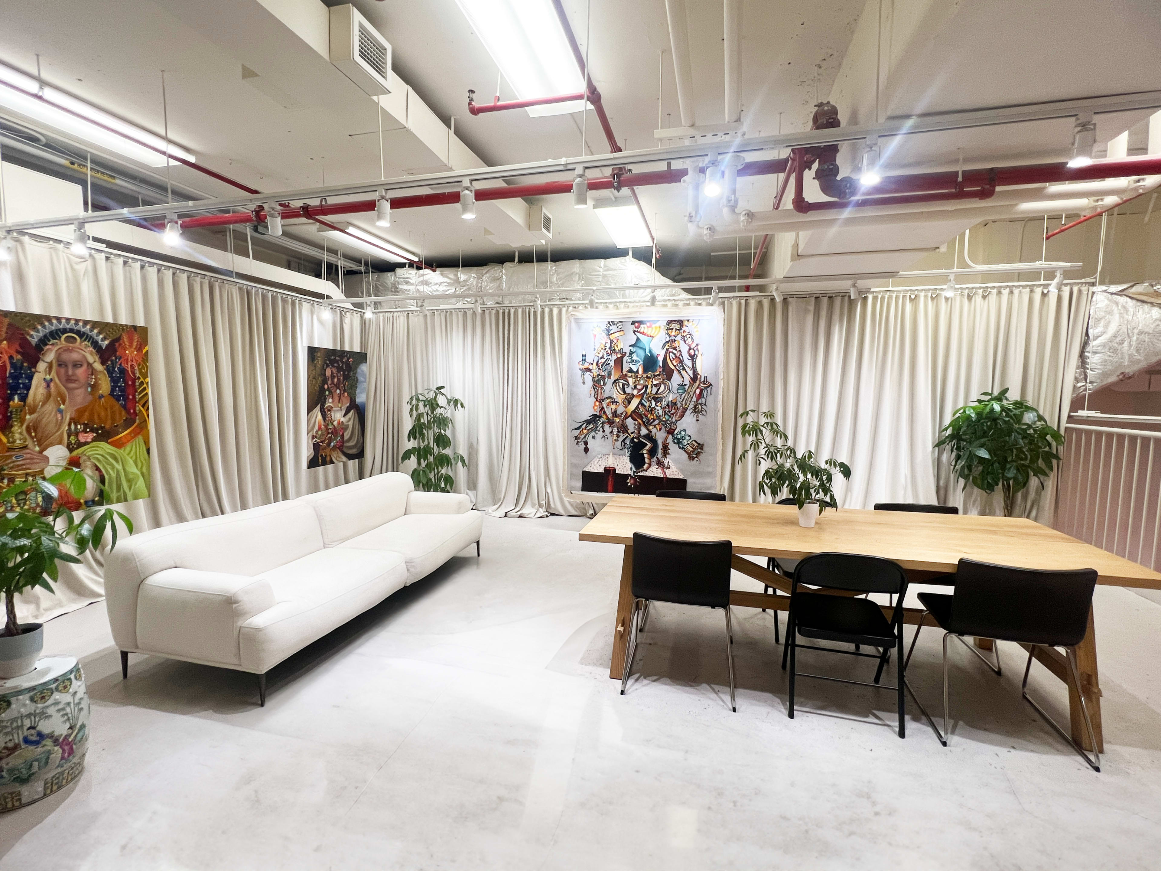 A spacious room with a white sofa, a wooden dining table with black chairs, potted plants, and colorful artwork on the walls, enclosed by curtains.