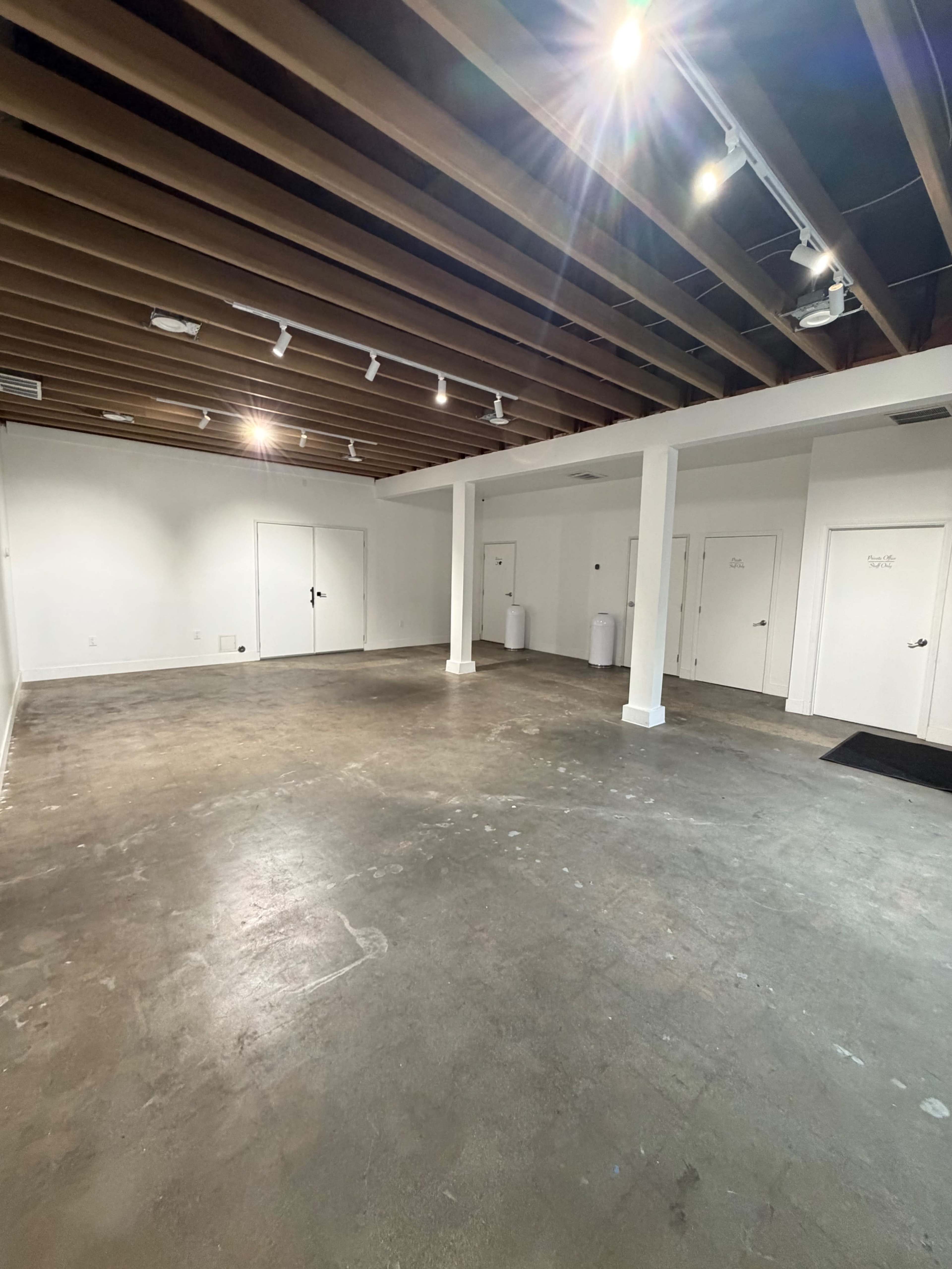 The image shows an empty room with polished concrete flooring, exposed wooden beams on the ceiling, and several closed doors along the walls.