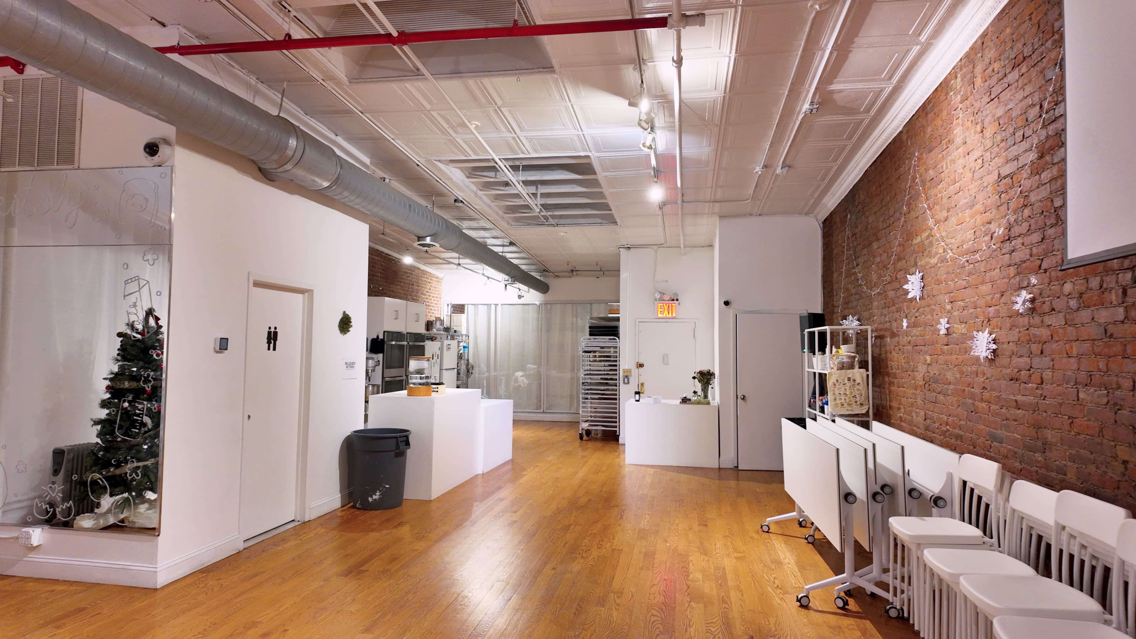 The image shows a brightly lit indoor space with wooden flooring, exposed brick walls, and a kitchen area featuring white countertops and a row of chairs along one side.