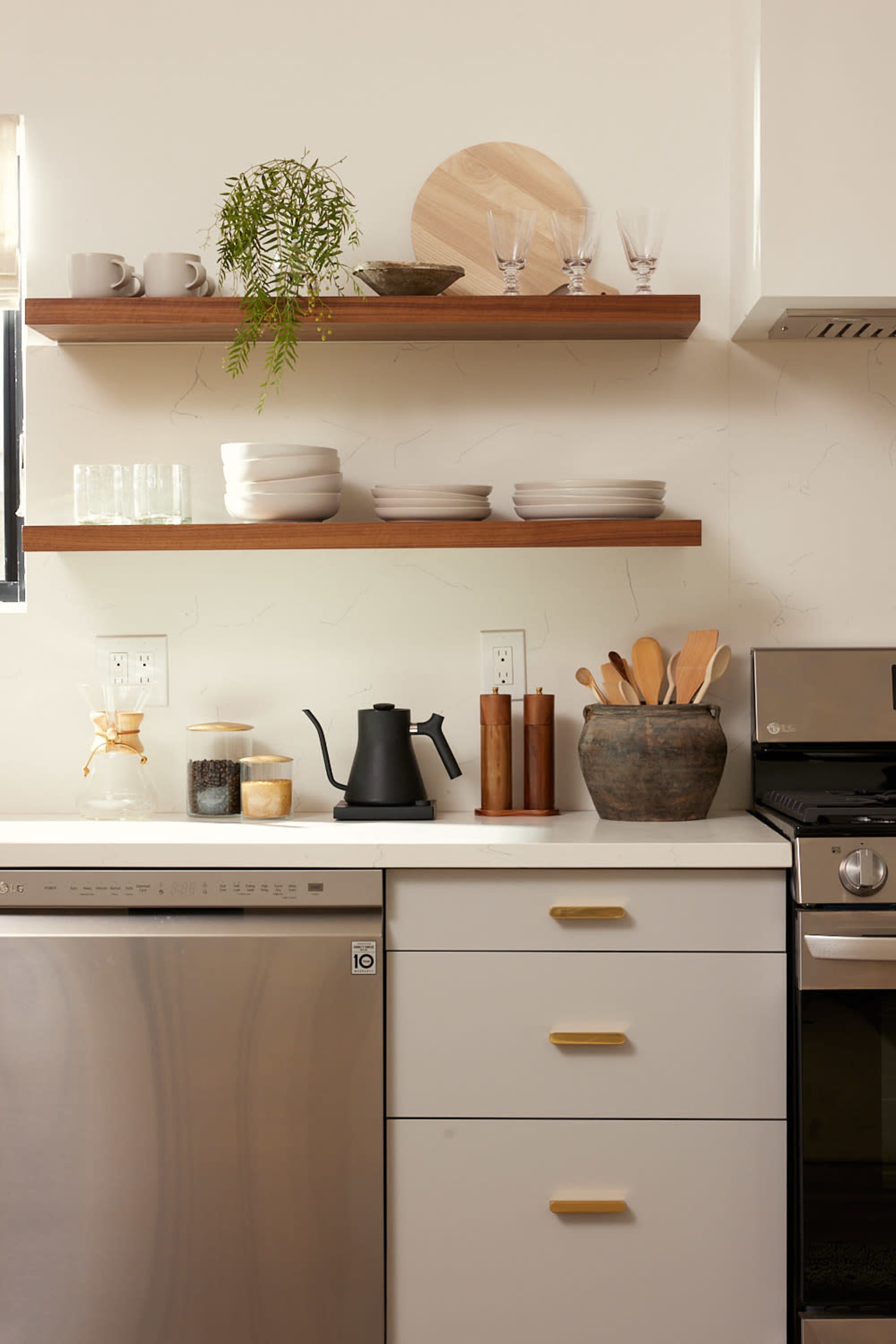 A modern kitchen with open wooden shelves displaying dishware and decorative items above a stainless steel dishwasher and an oven.