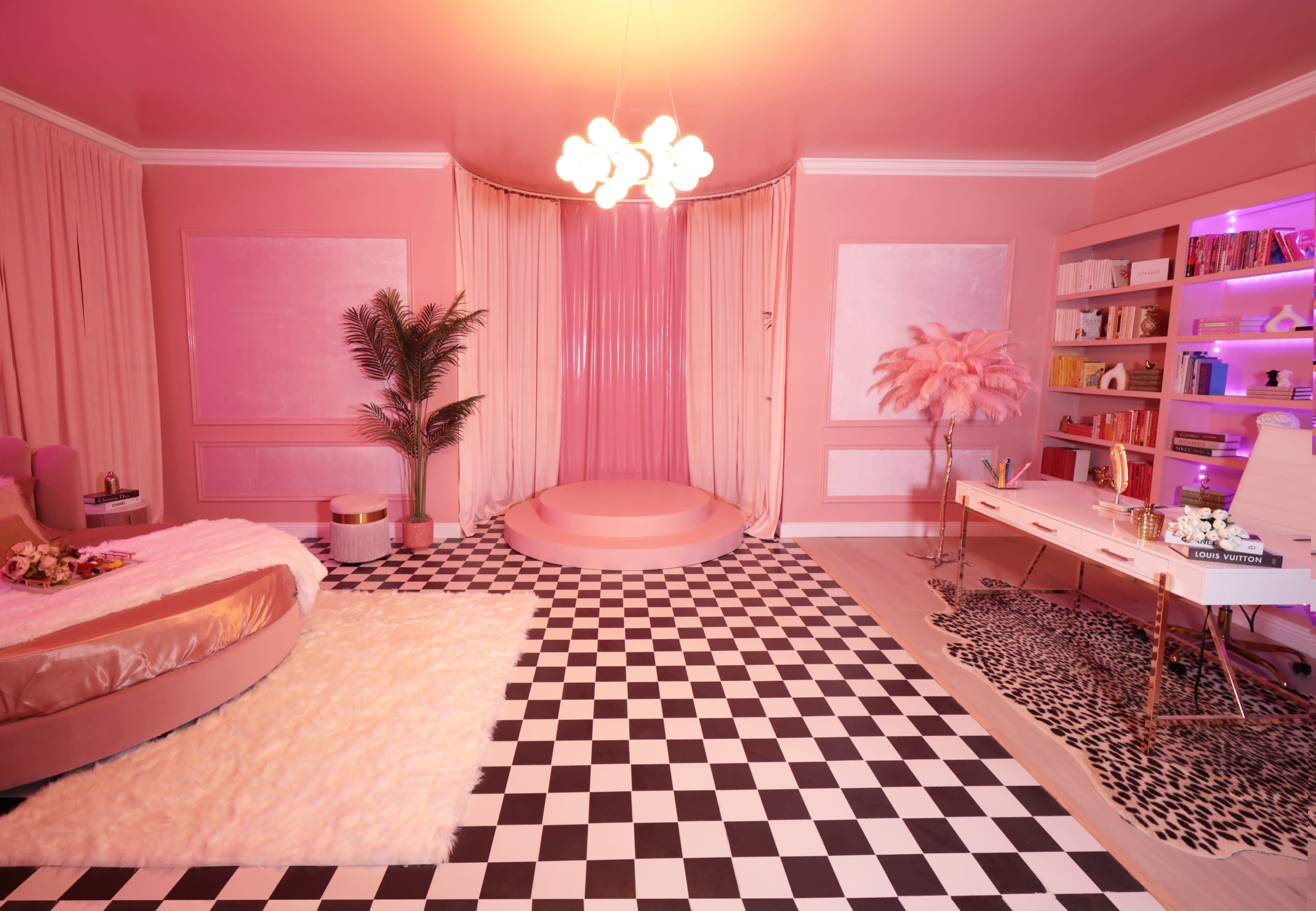 A pink-themed room features a circular stage, a checkered floor, and a white desk with shelving filled with books and decorative items.