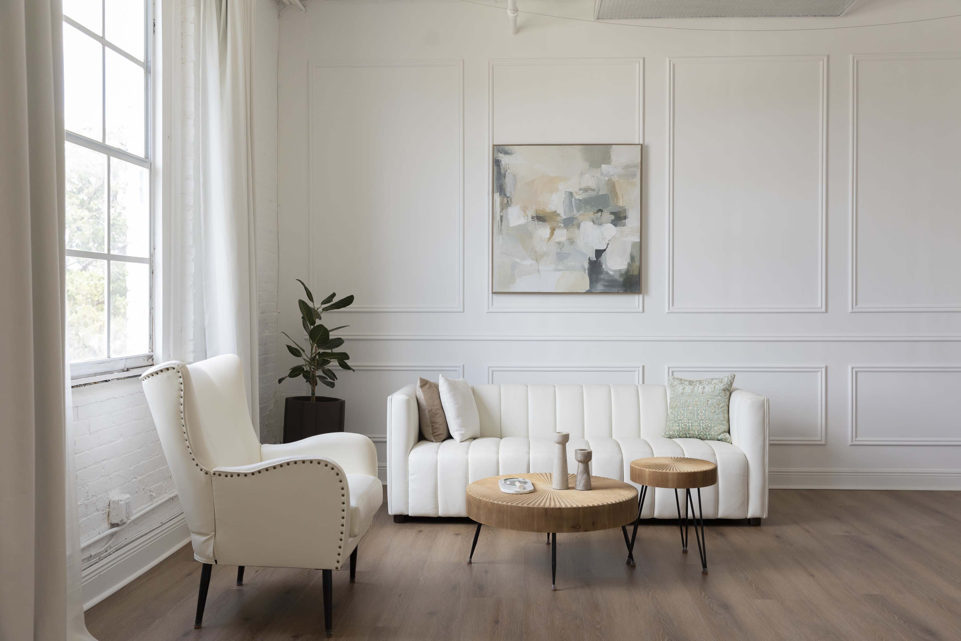 The image shows a modern living room with a white sofa, two wooden coffee tables, and a potted plant beside a large window.