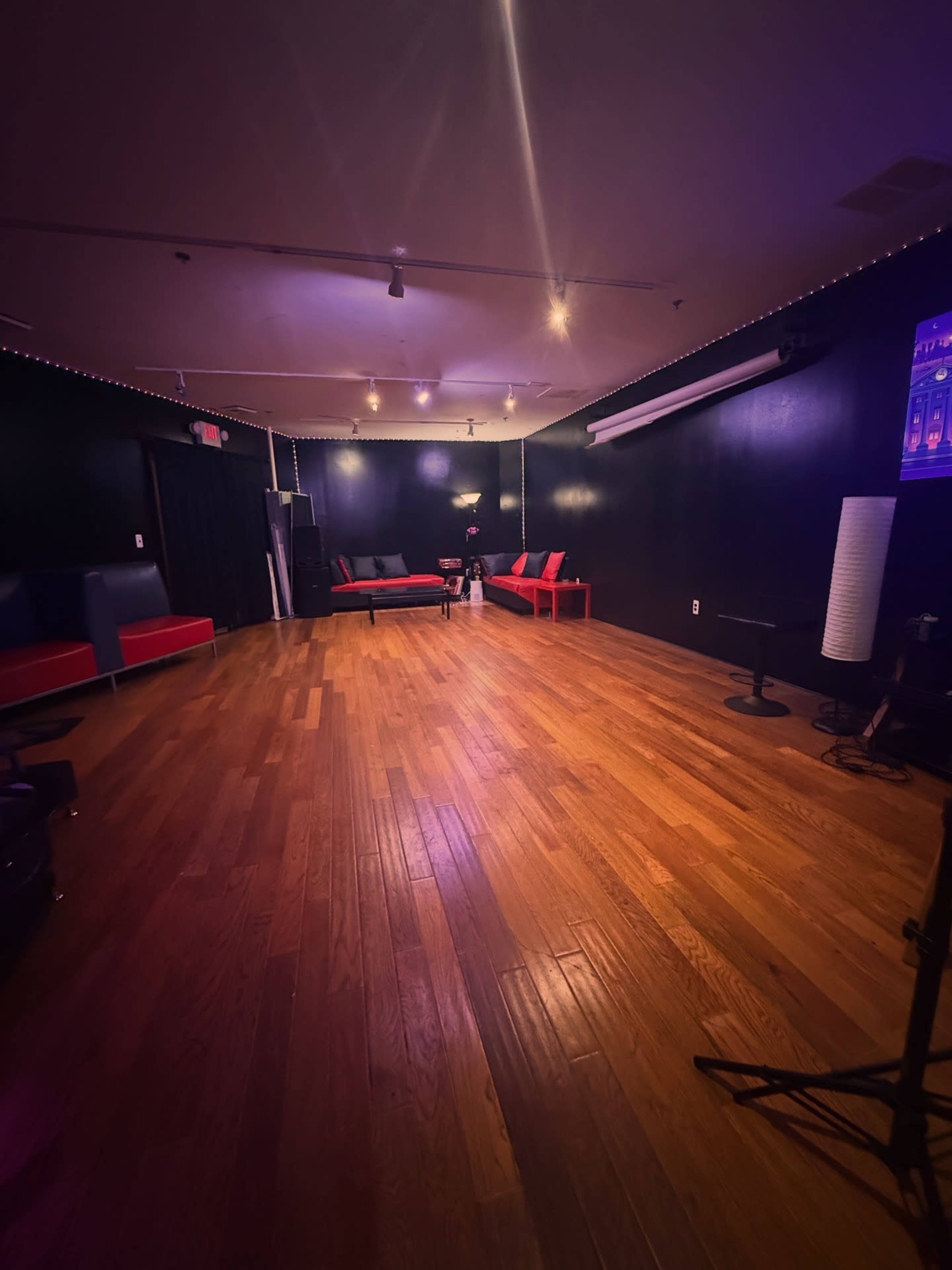 The image shows a spacious, dimly lit room with hardwood flooring, featuring red and black seating arrangements along the walls.