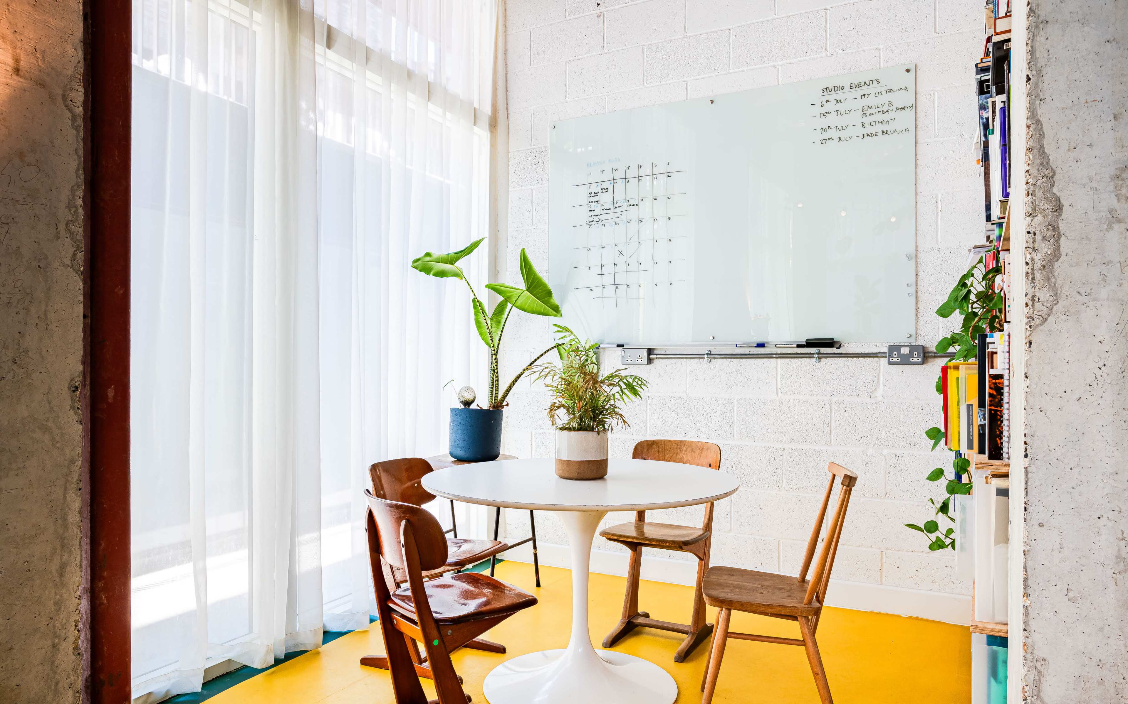 A bright room features a round white table with four wooden chairs, a potted plant, and a whiteboard on the wall, illuminated by natural light through sheer curtains.