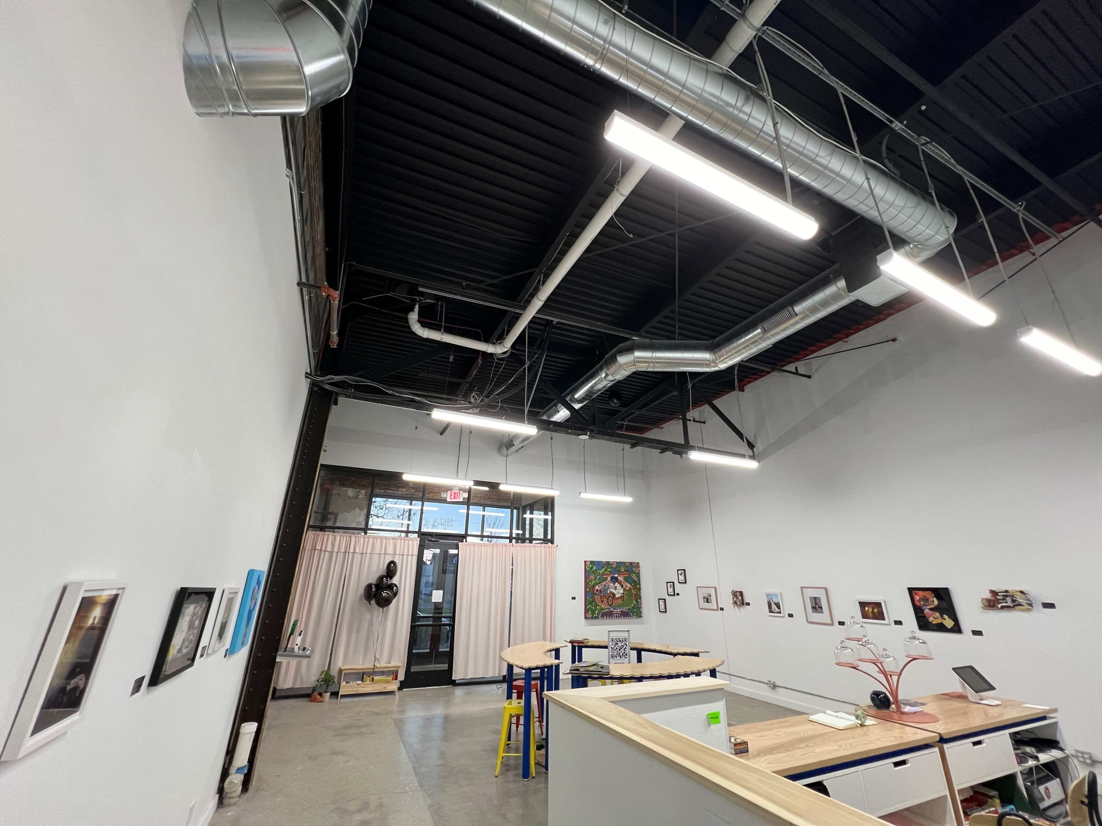 The image shows a spacious interior office or studio with exposed ductwork, large windows, and various framed artworks on the walls.