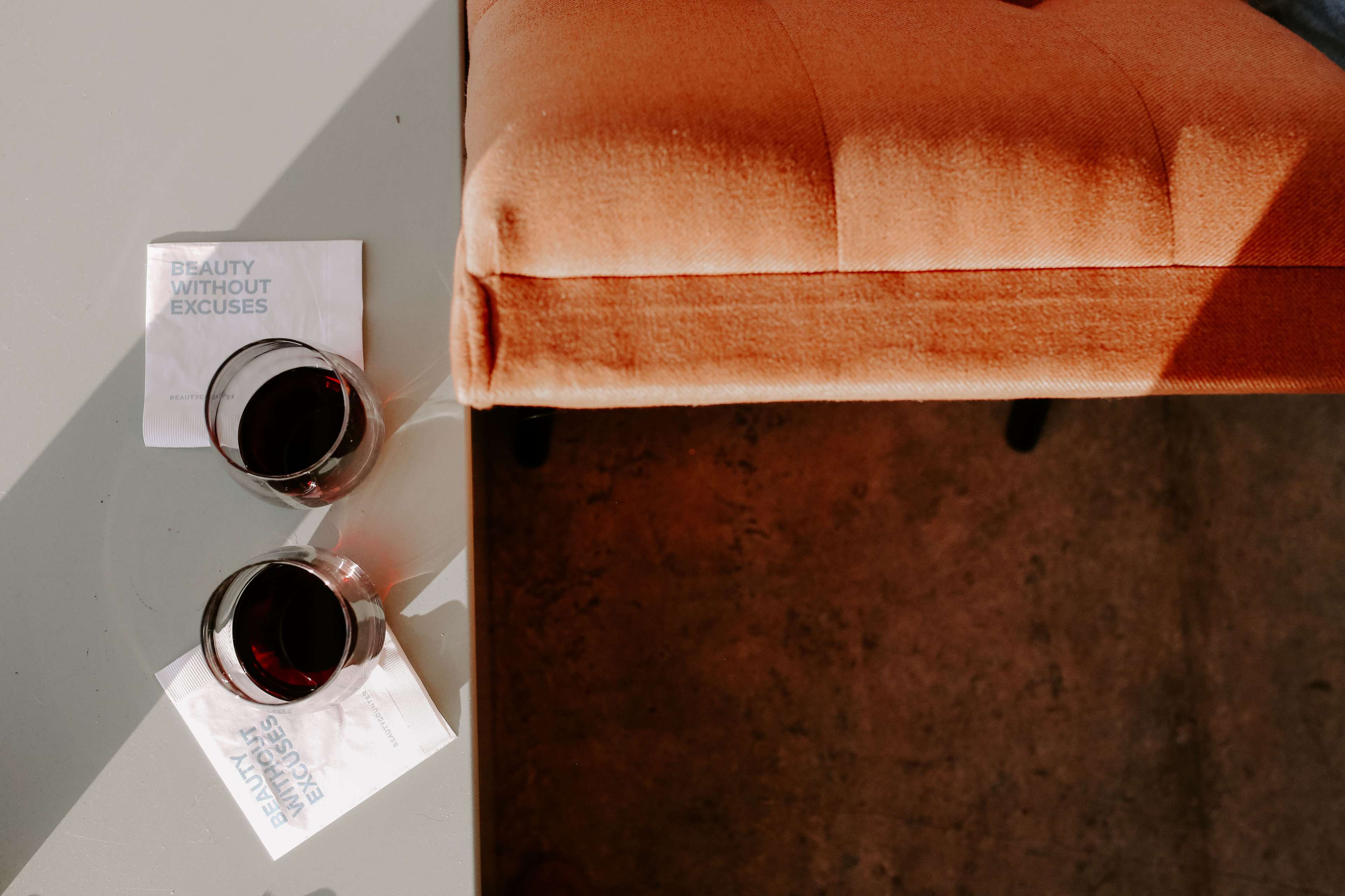 Two glasses of red wine sit on white napkins next to a tufted orange chair on a table.