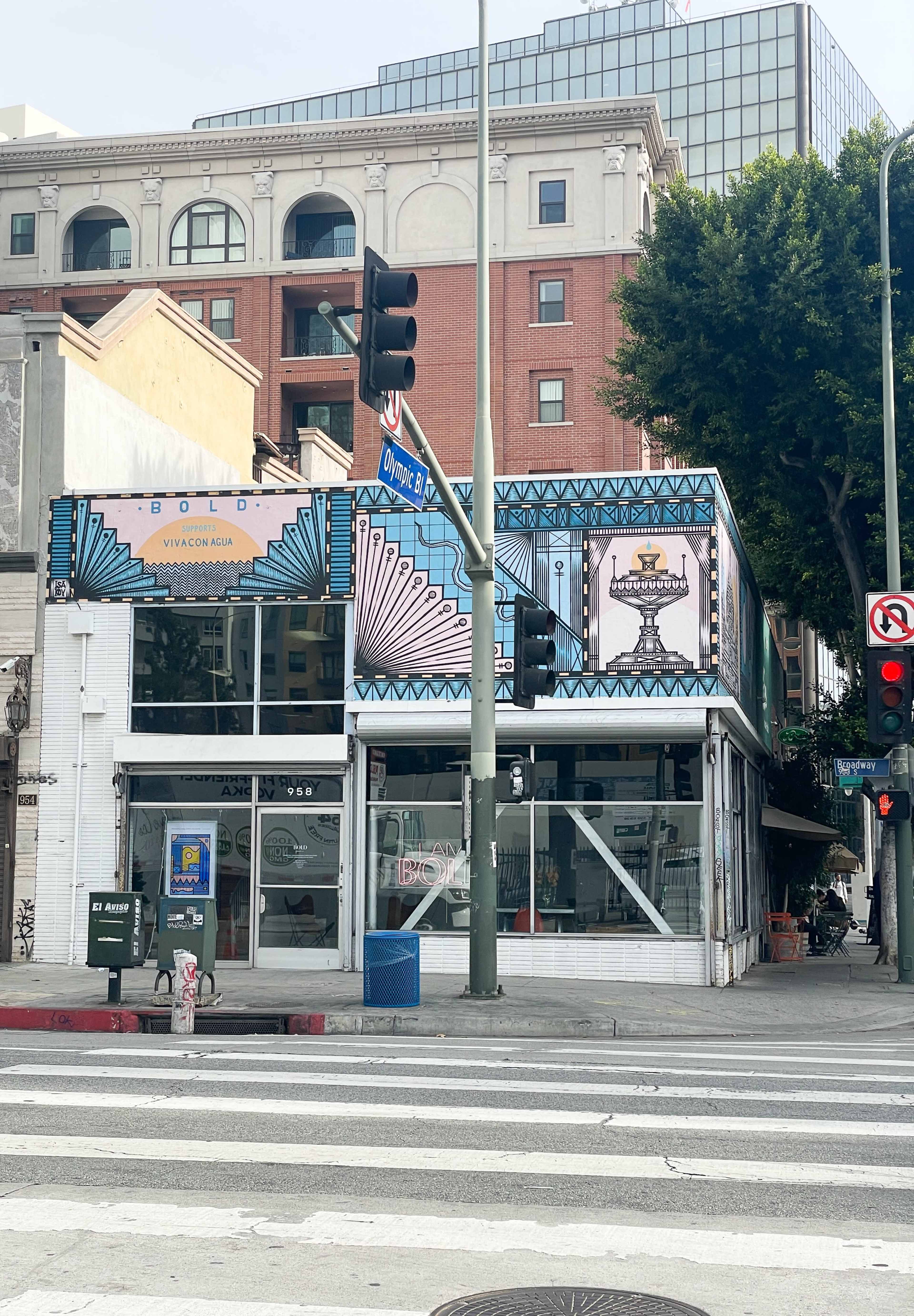 A building with a colorful façade and a large mural is located at the intersection of Olympic Boulevard and another street, with traffic lights visible.