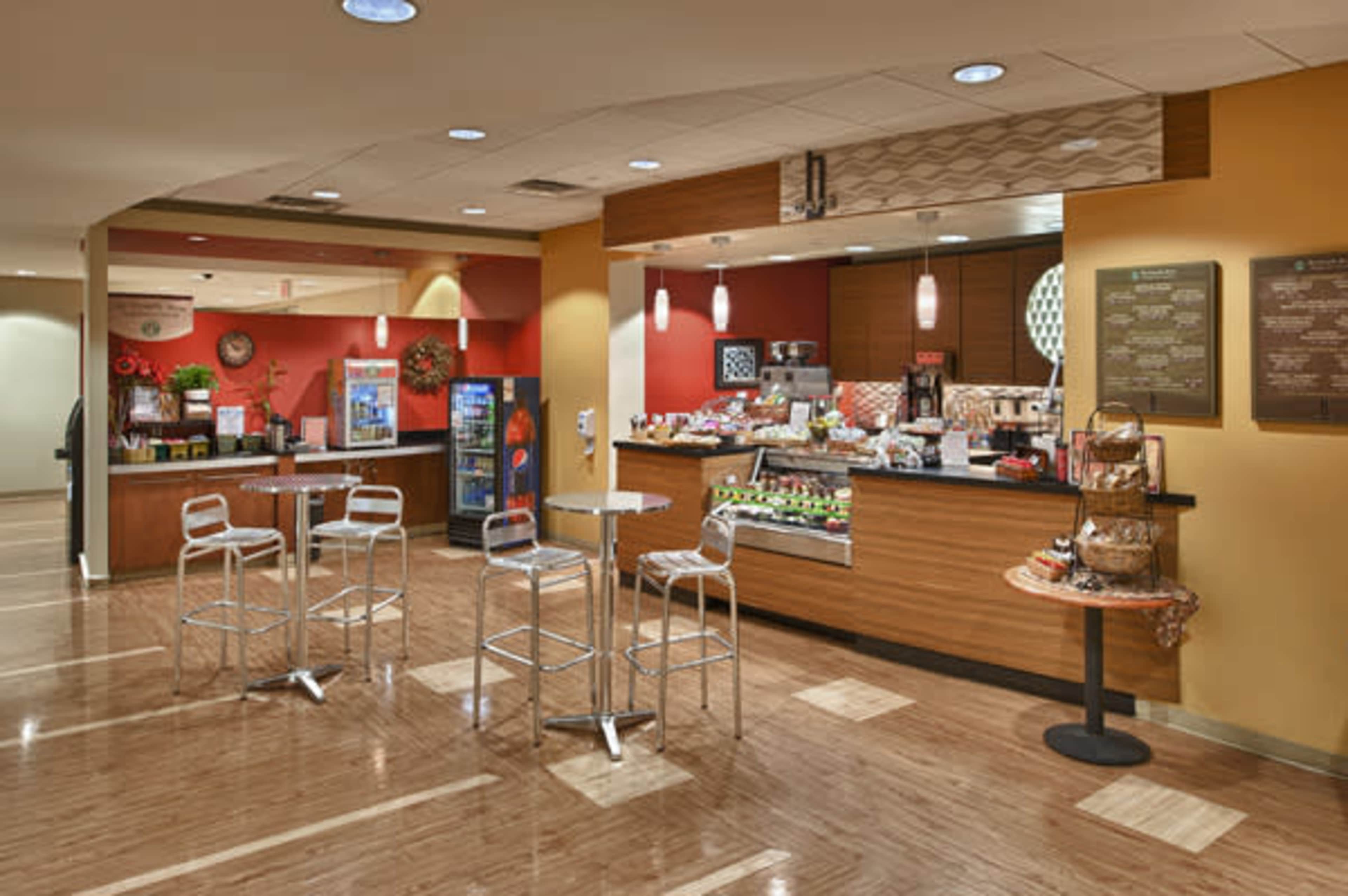 The image shows a modern café with a seating area and a service counter displaying food and beverage options.