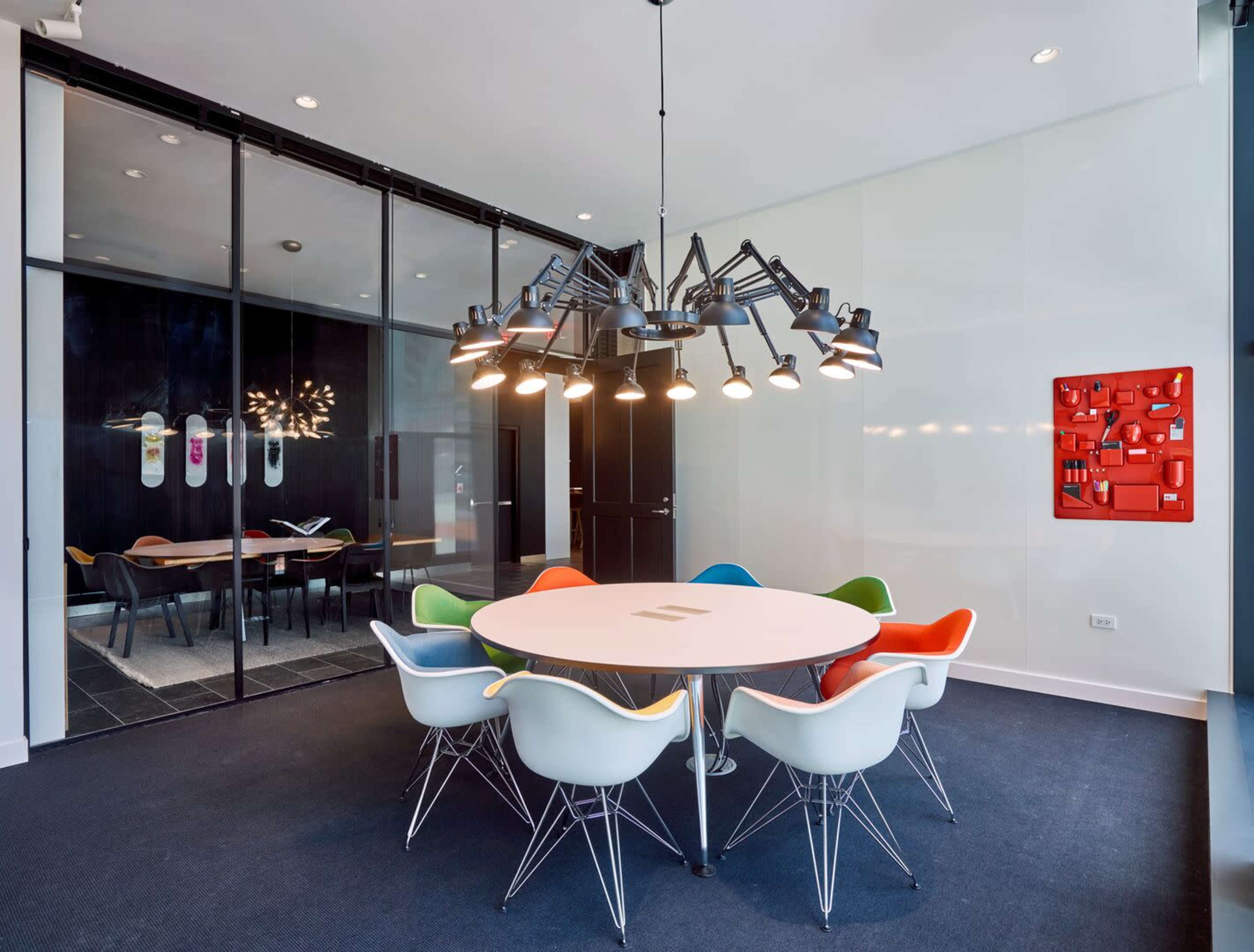 A modern conference room features a round table surrounded by colorful chairs and a large chandelier, with a glass wall revealing another meeting area.
