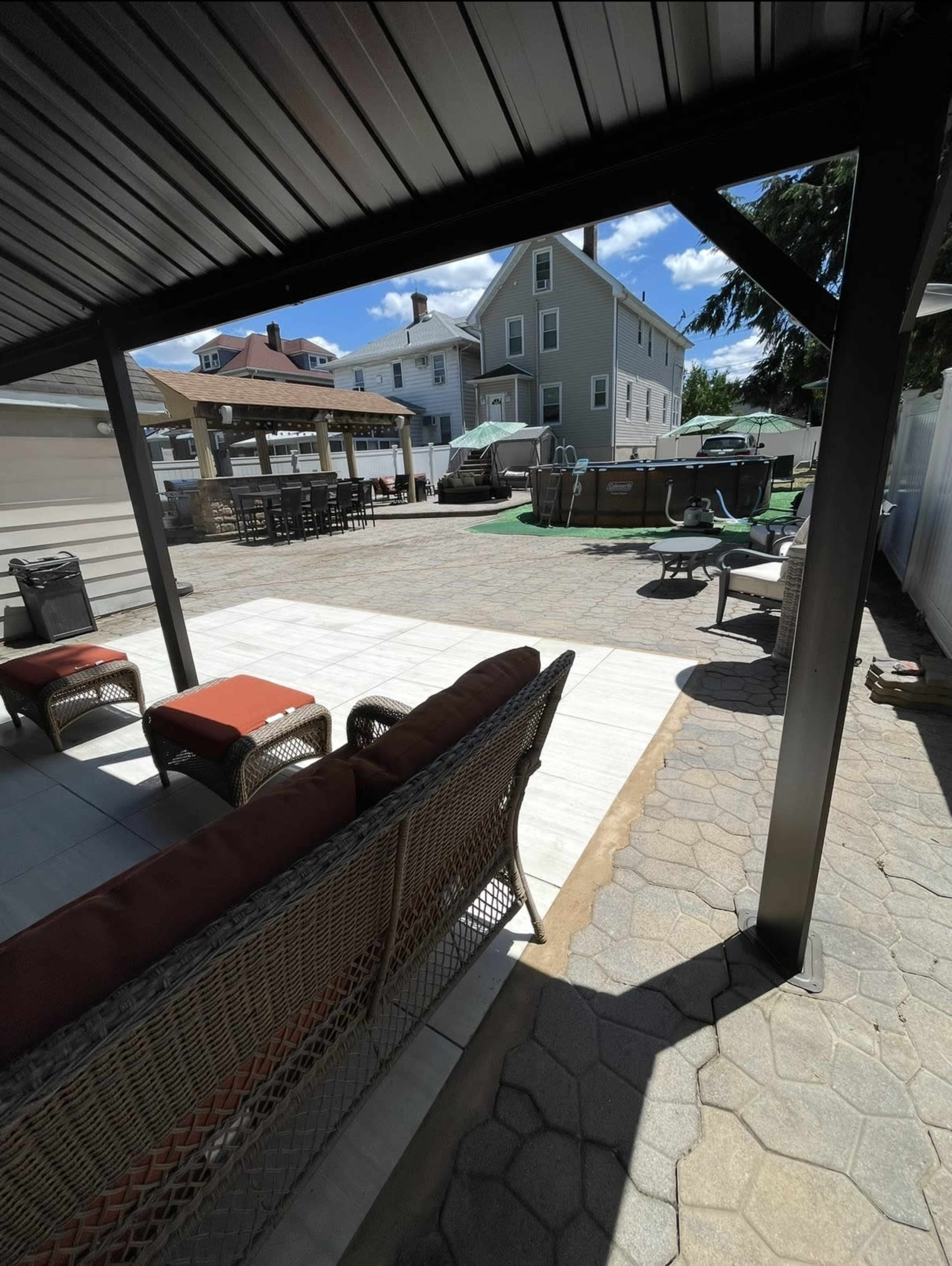 The image shows a patio area with a seating arrangement under a roof, overlooking a spacious outdoor space featuring a grassy area, parked vehicles, and shaded tables.
