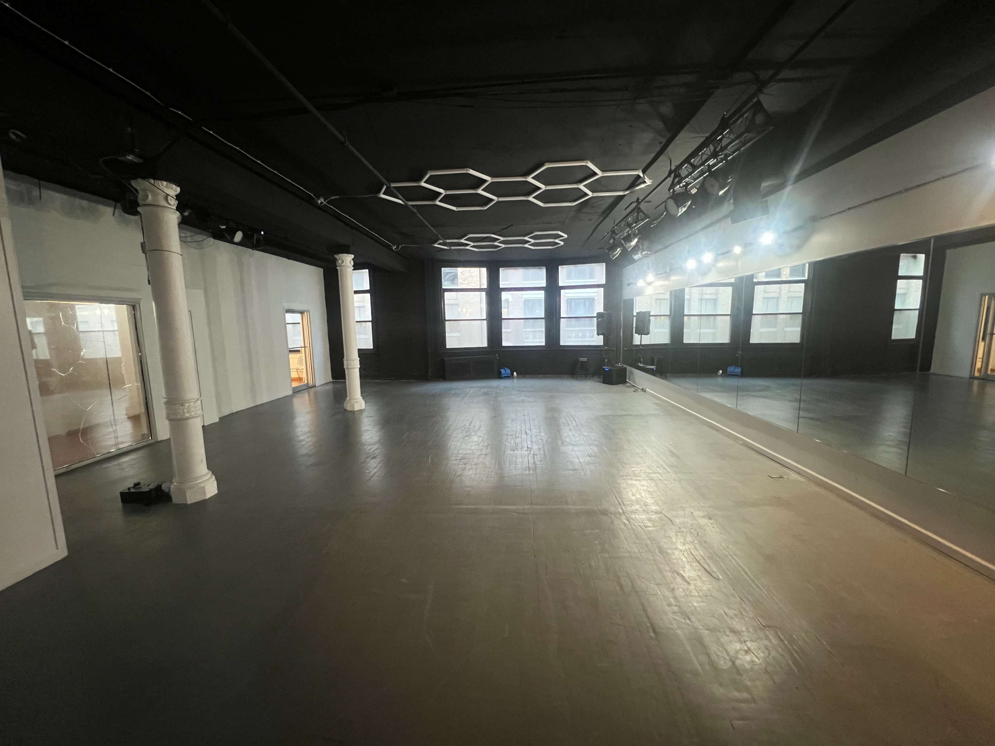 The image shows a large, empty studio space with hardwood floors, columns, a mirrored wall, and overhead lighting.