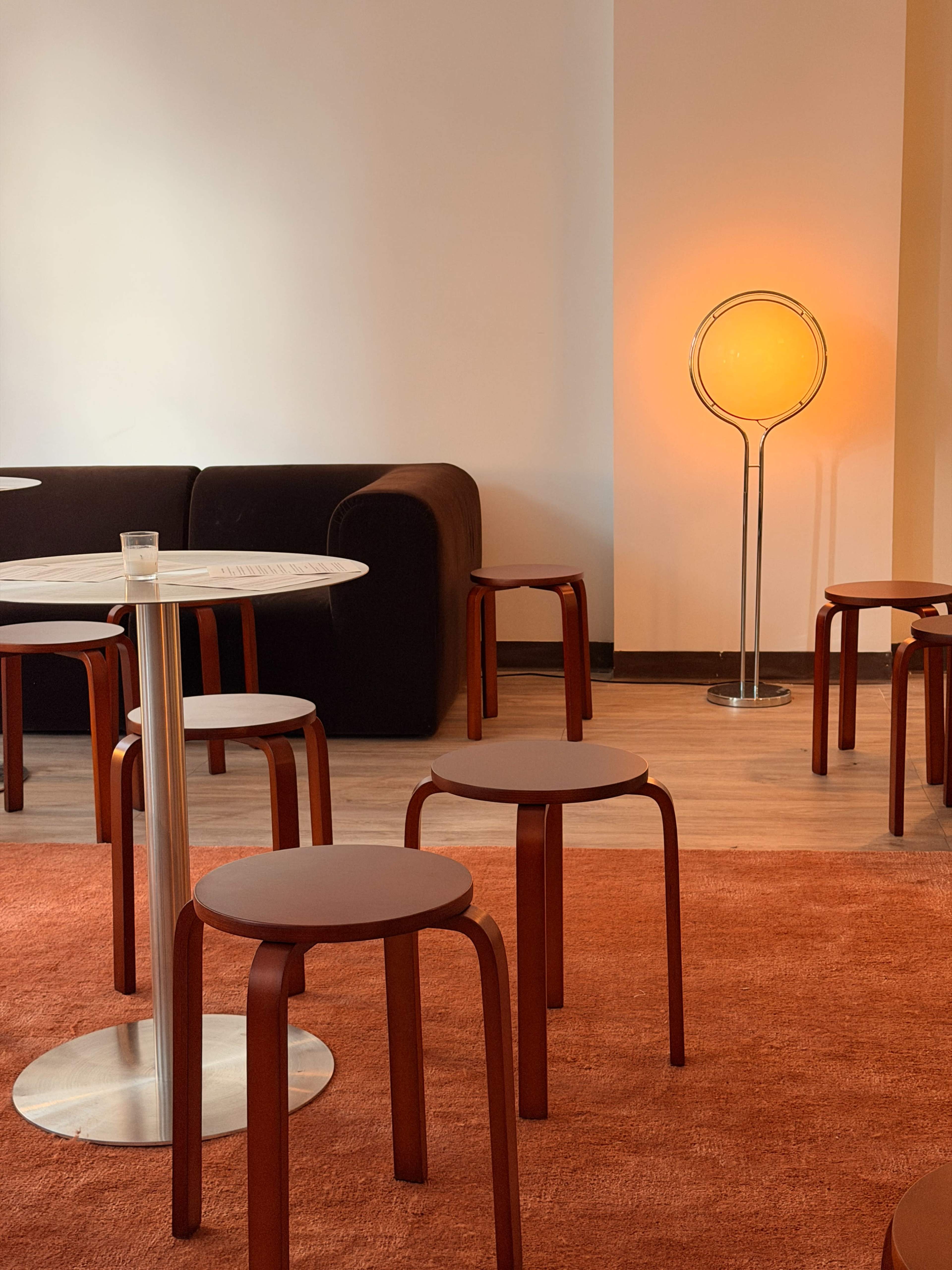 A minimalist café scene features a round table, several red stools, a black sofa, and an orange lamp in the corner.