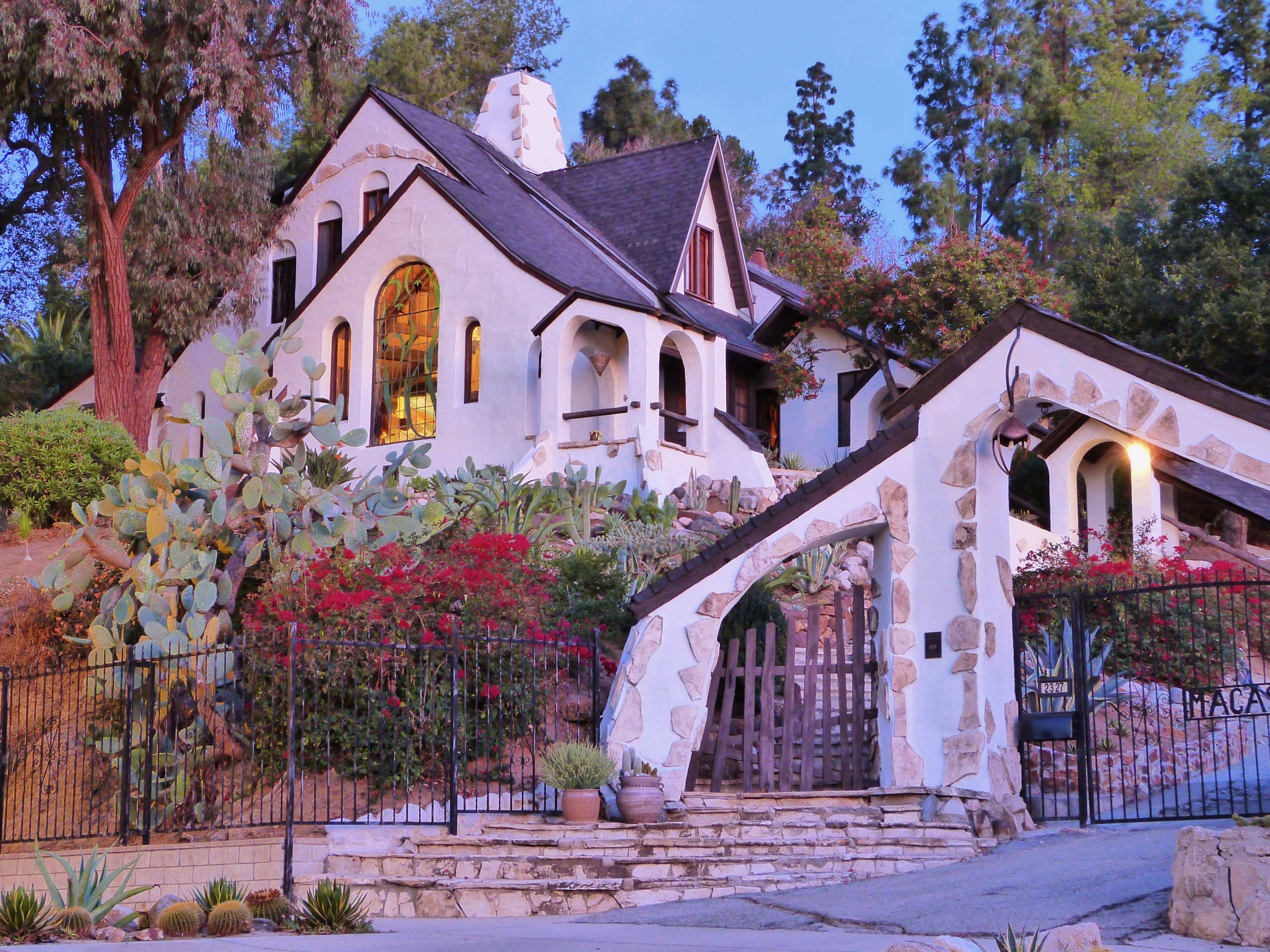 A white, textured house with a steep roof and arched windows is surrounded by lush greenery and colorful flowers.