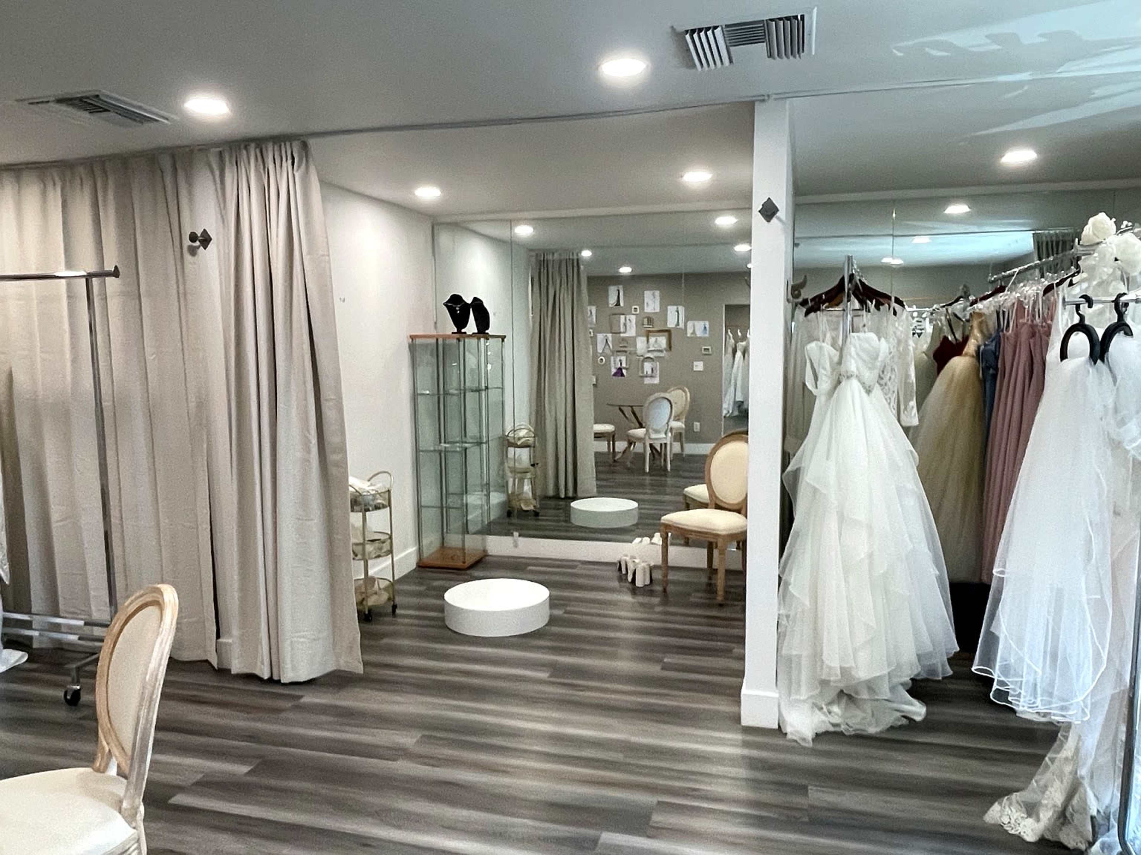 The image shows a bridal boutique with wedding dresses displayed on racks, a seating area with a mirror, and a glass display case.