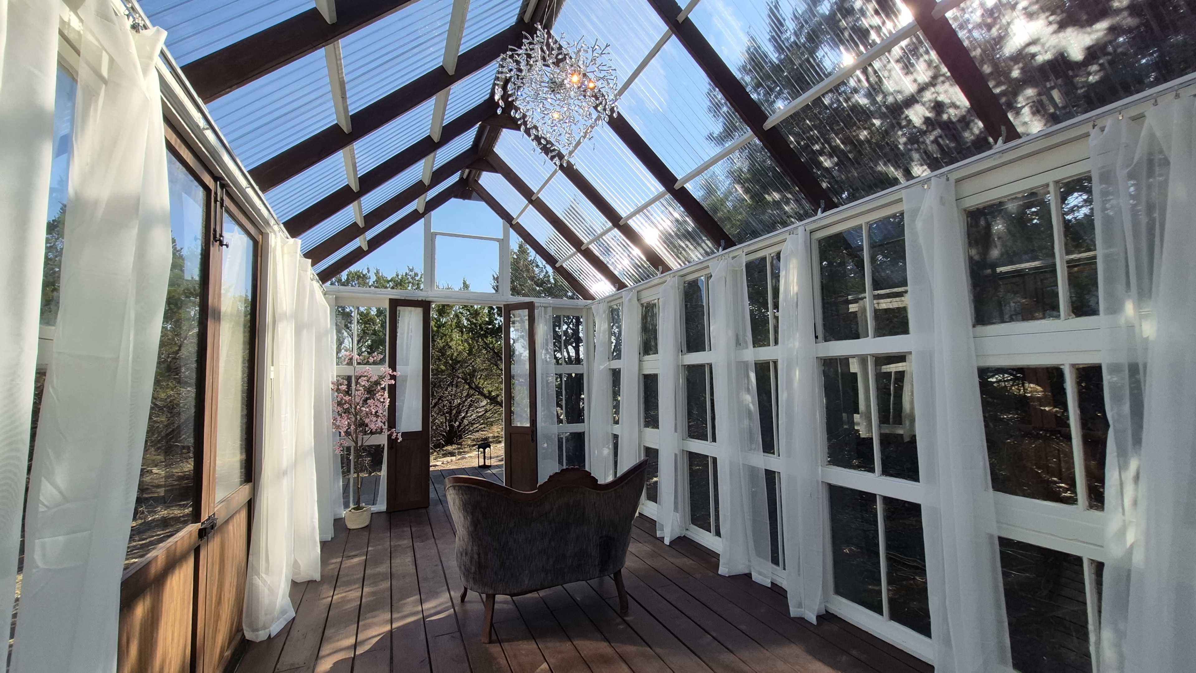 The image shows a sunlit glass greenhouse with wooden flooring, featuring large windows, white drapes, and an elegant chair in the center.
