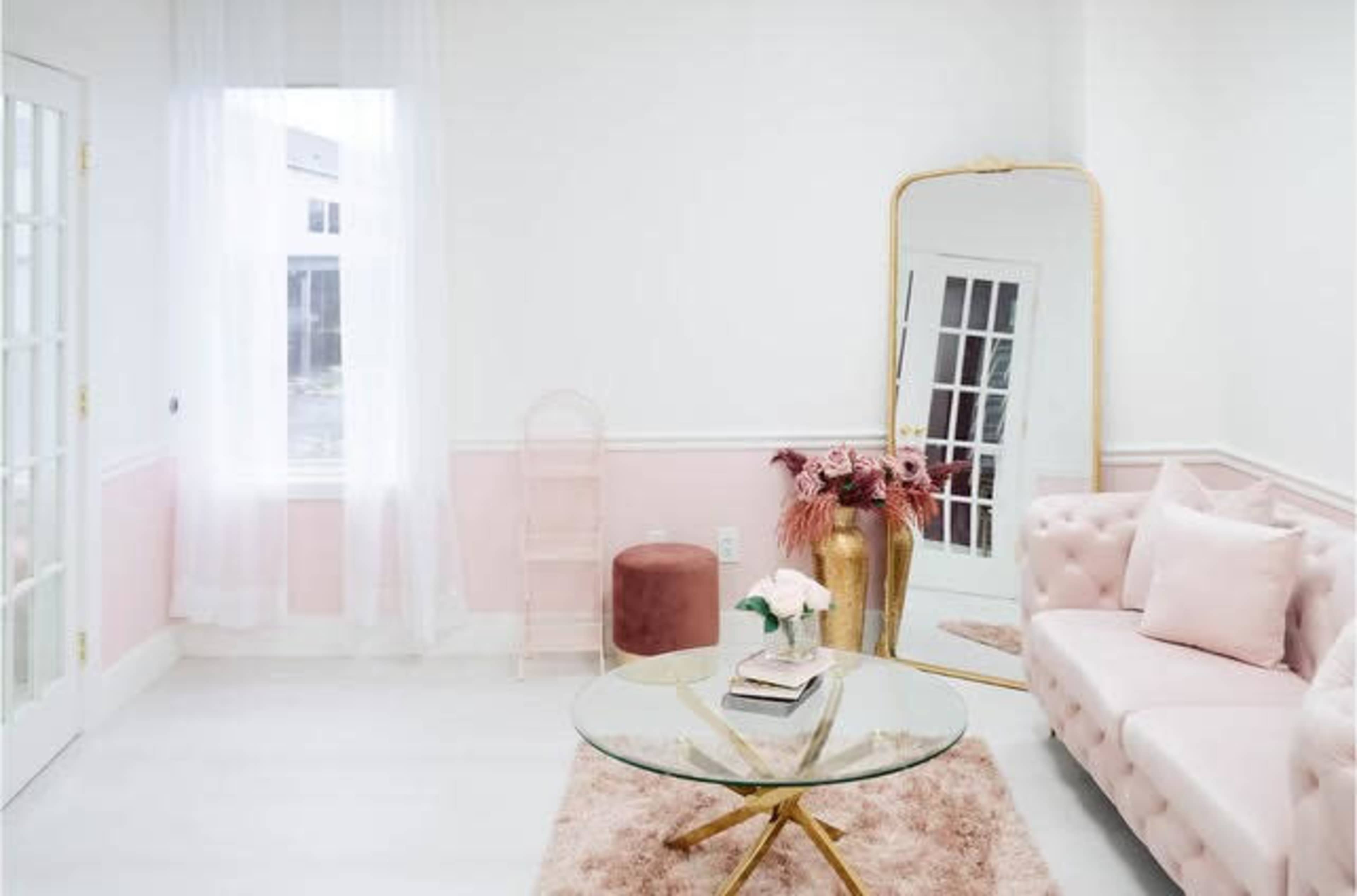 The room features a light-colored décor with a pink tufted sofa, a glass coffee table, a large mirror, and decorative plants.
