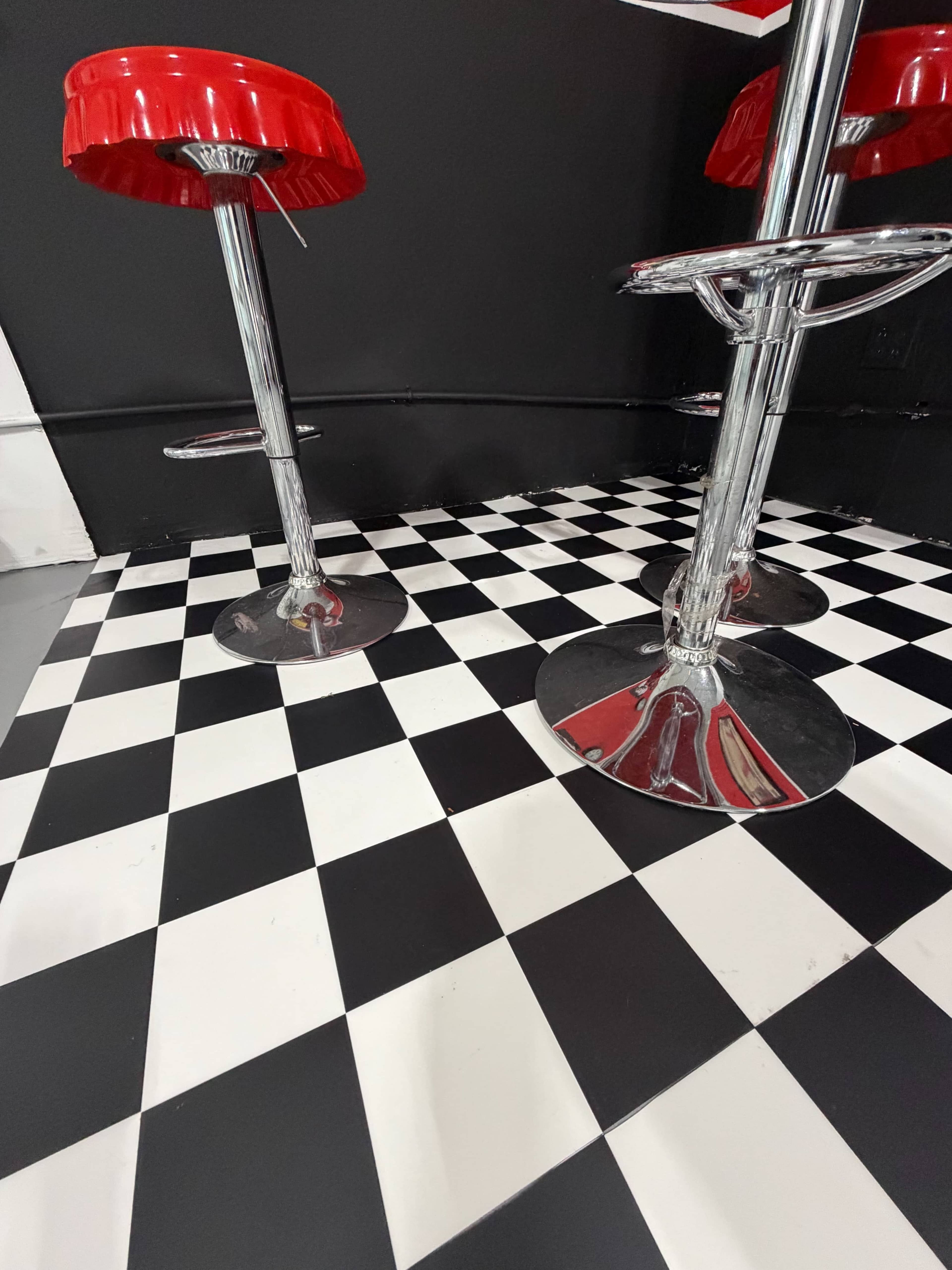 The image shows a corner of a room with black and white checkered flooring and two chrome bar stools with red seats.