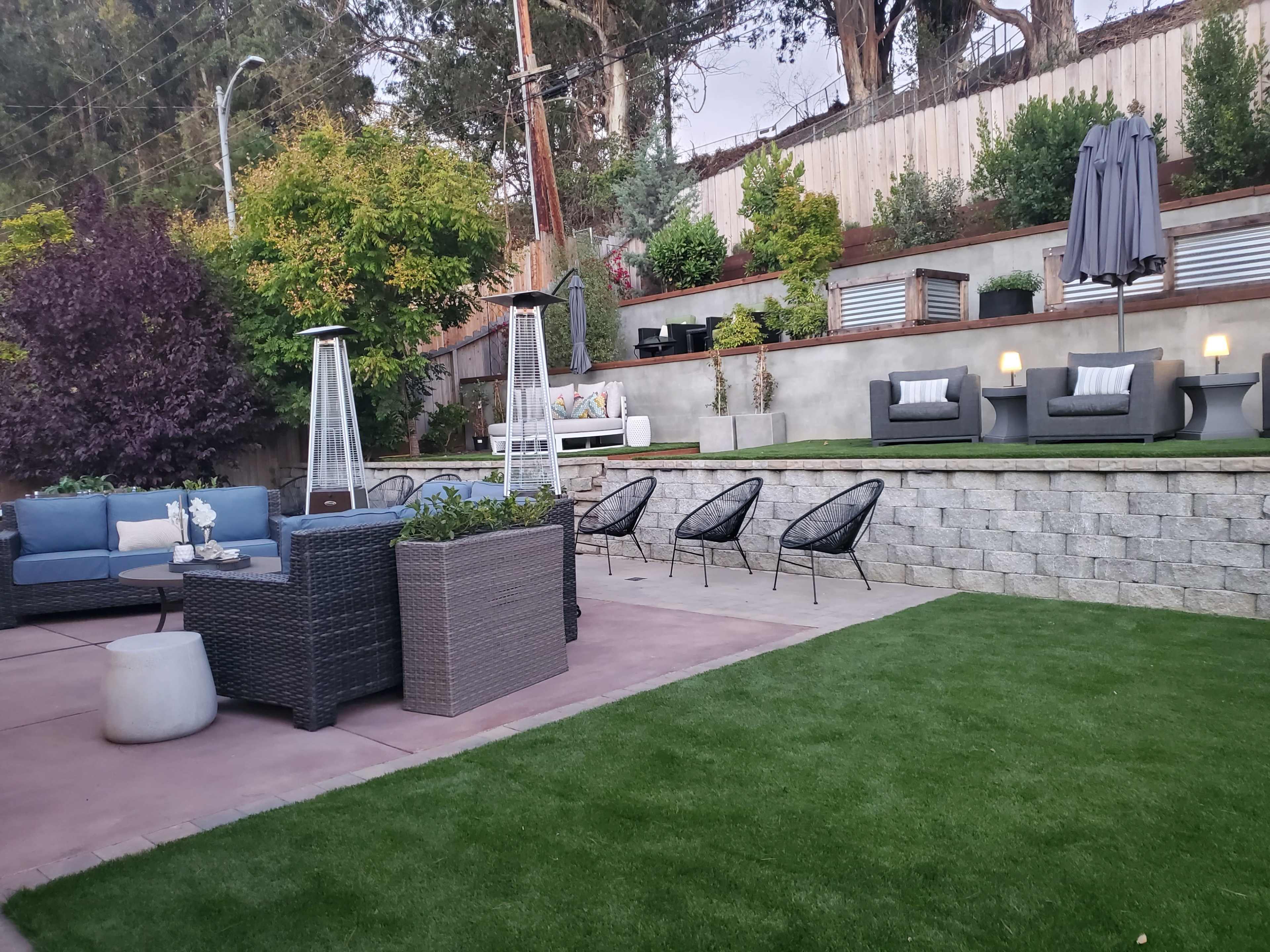 A landscaped backyard with seating areas, including a sofa set, lounge chairs, and a fire pit, bordered by greenery and stone walls.