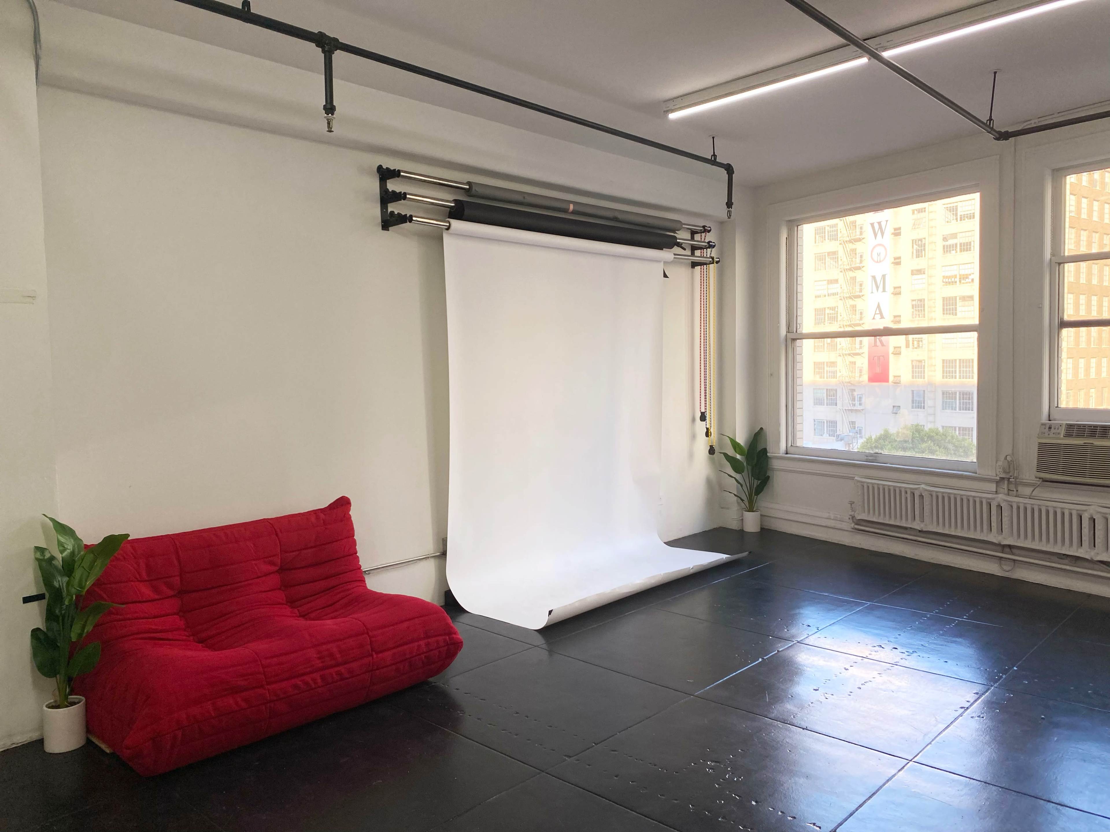 A minimalist studio space with a red sofa, a white backdrop setup, and large windows letting in natural light.