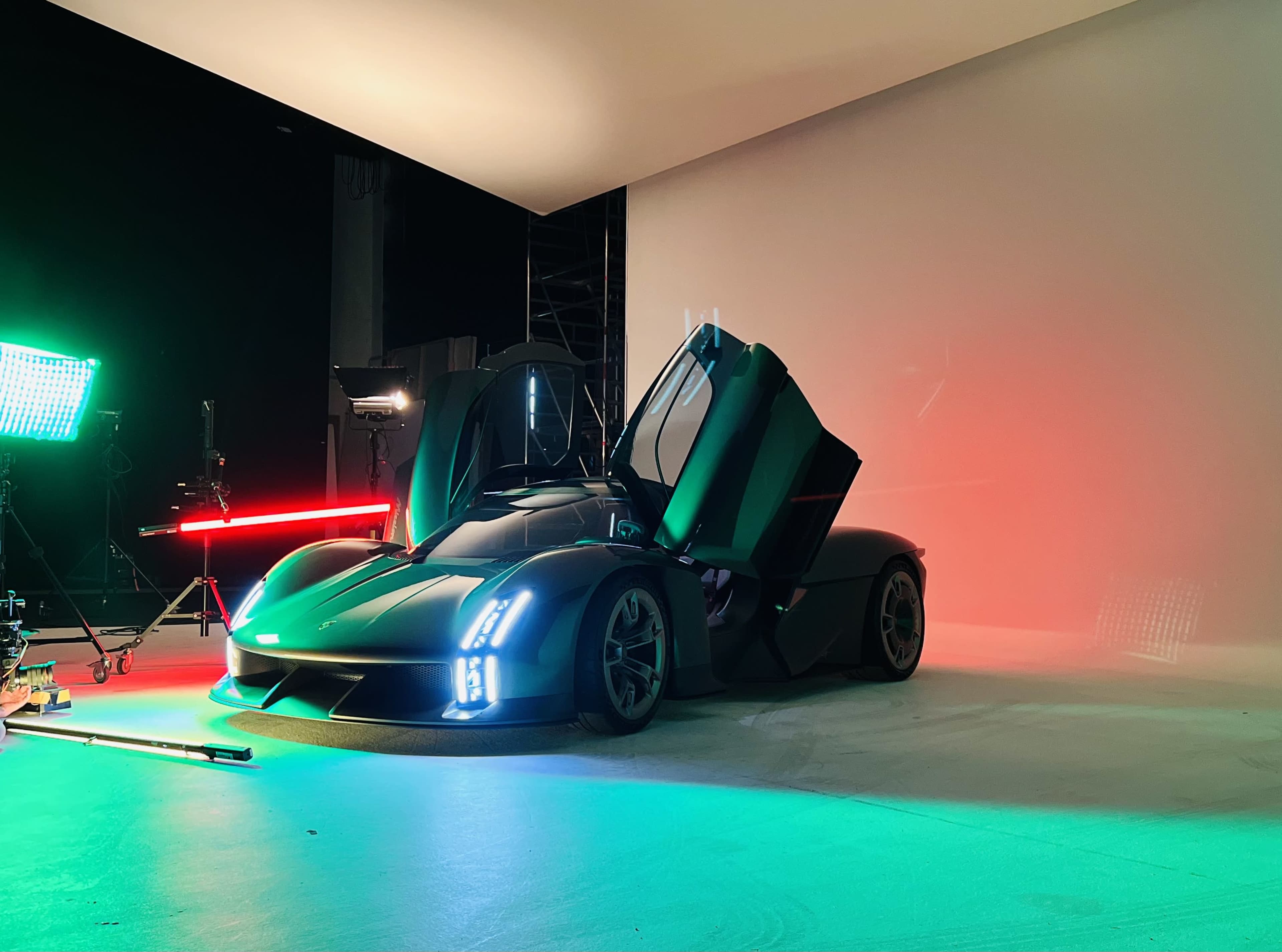 A sleek sports car with upward-opening doors is positioned on a stage surrounded by colorful lighting and studio equipment.
