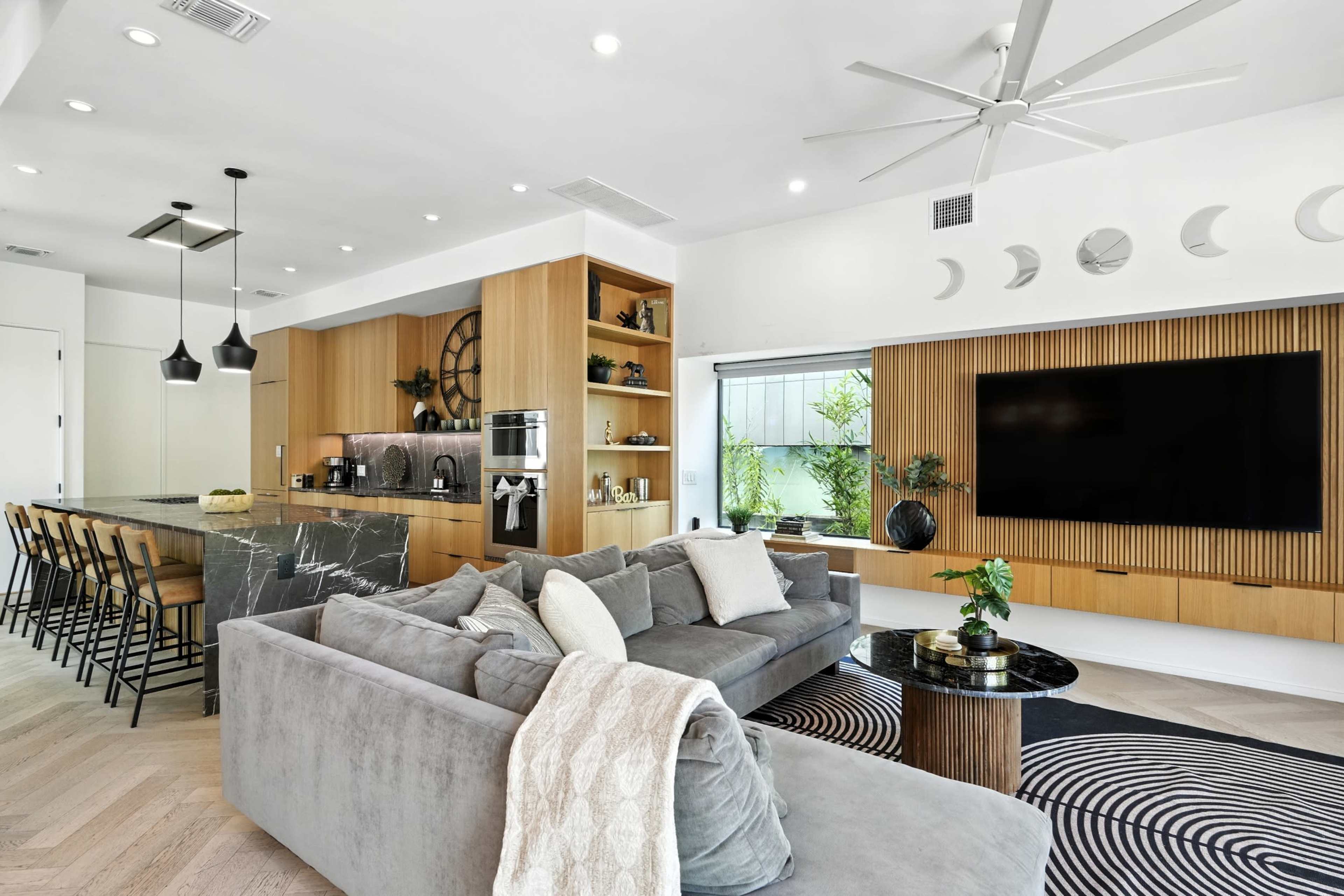 The image shows a modern living room with a large sofa, a round coffee table, and an open kitchen featuring a marble countertop and bar seating.