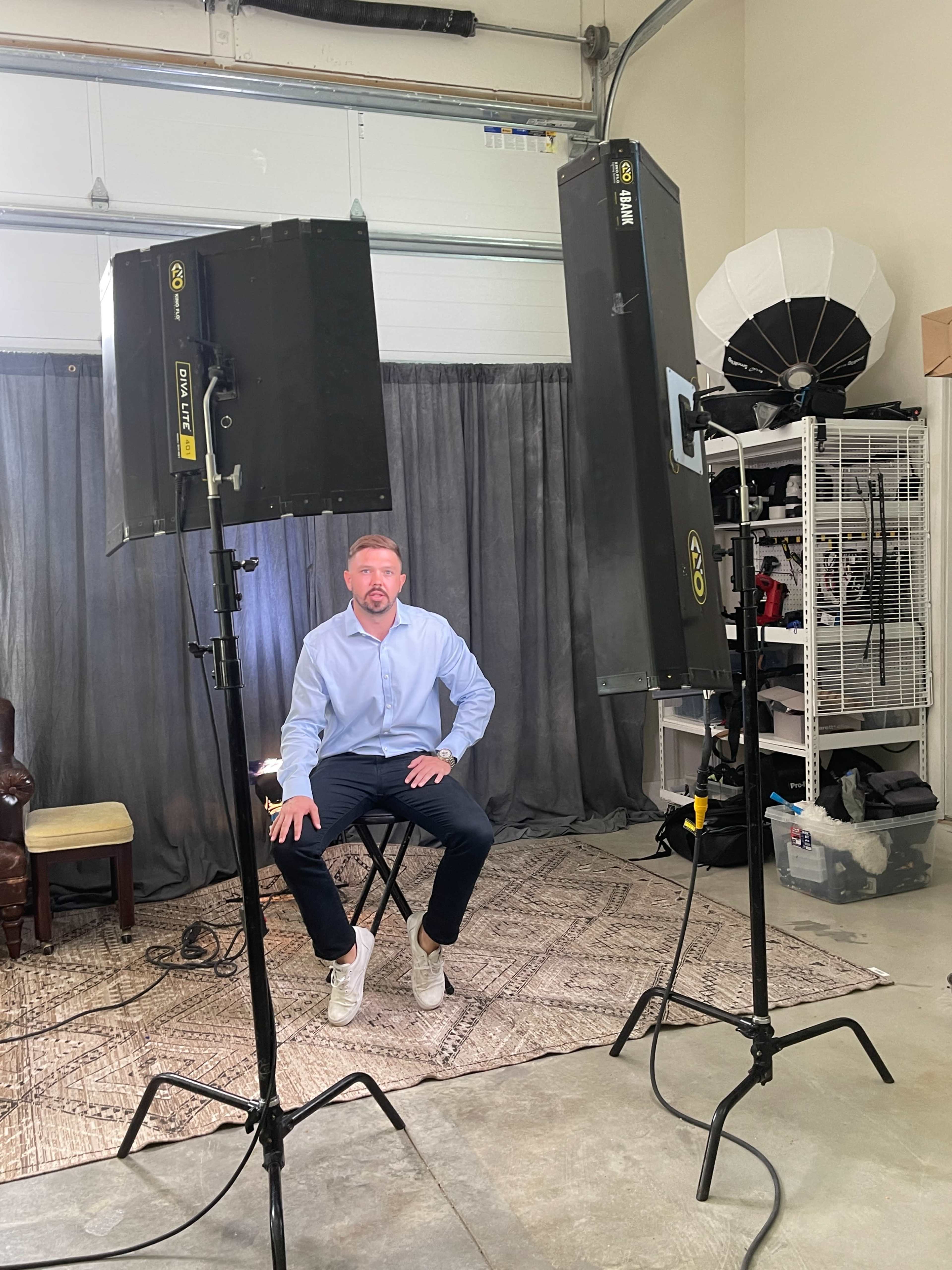 A man in a light blue shirt sits on a stool in front of two studio lights in a photography setting.